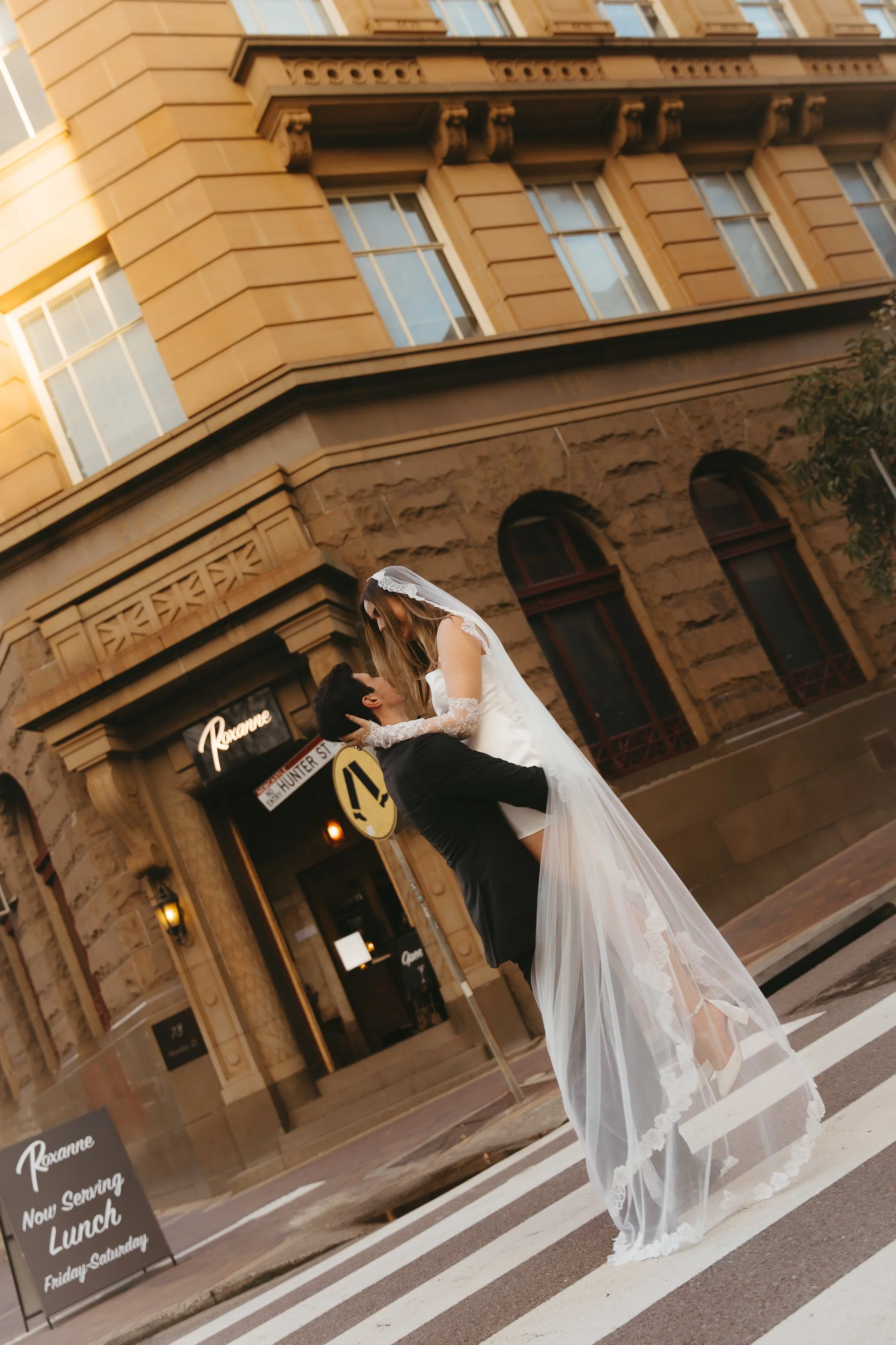 A bride and groom on a city street, with the groom lifting the bride while they look at each other. The bride is wearing a white wedding dress and veil, and the groom is in a black suit. Behind them is an old building with large windows and a restaur