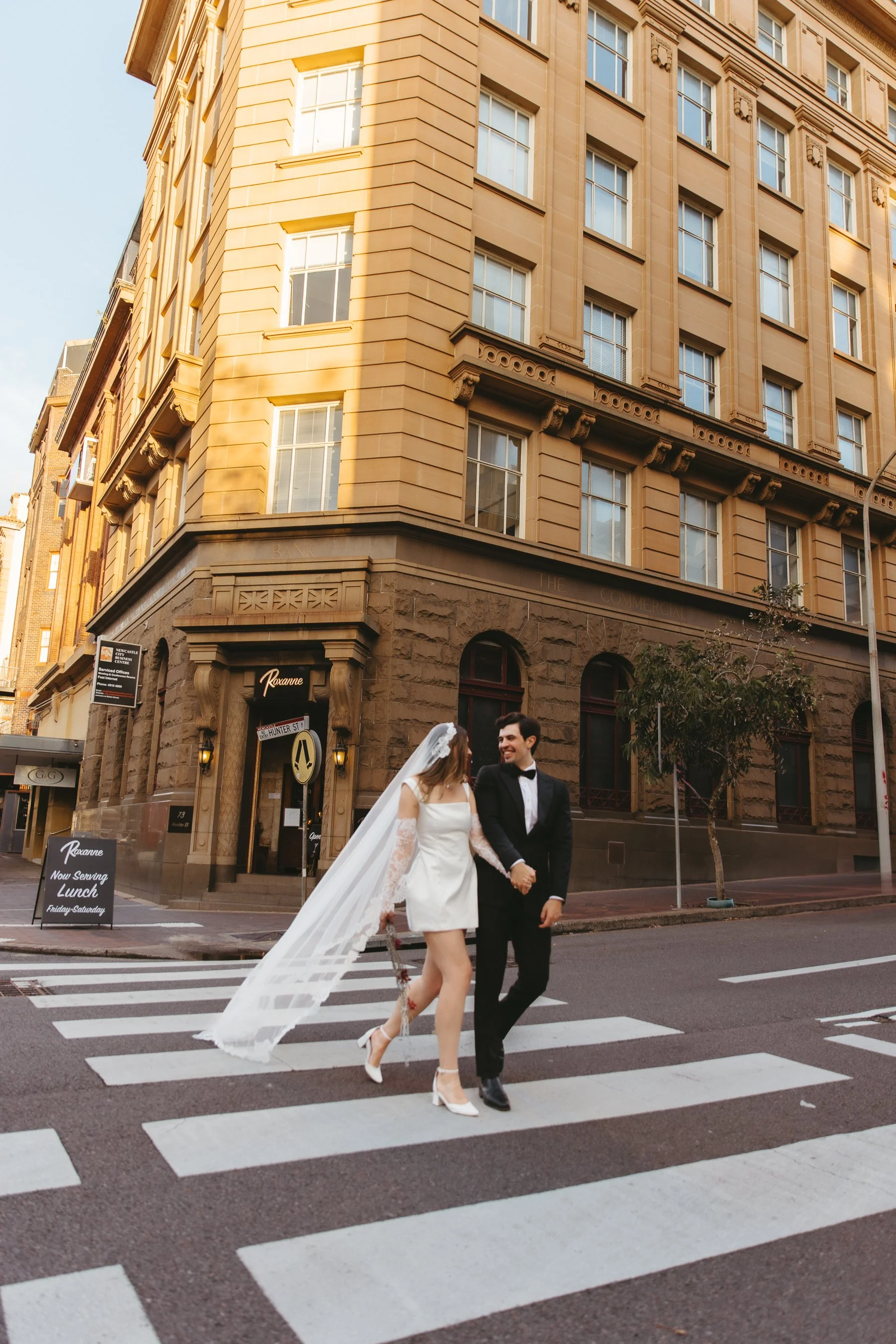 A bride and groom walking hand-in-hand across a city crosswalk. The bride is wearing a white dress with a veil and lace sleeves, and white high heels. The groom is dressed in a black tuxedo with a bow tie. They are smiling and looking at each other, 