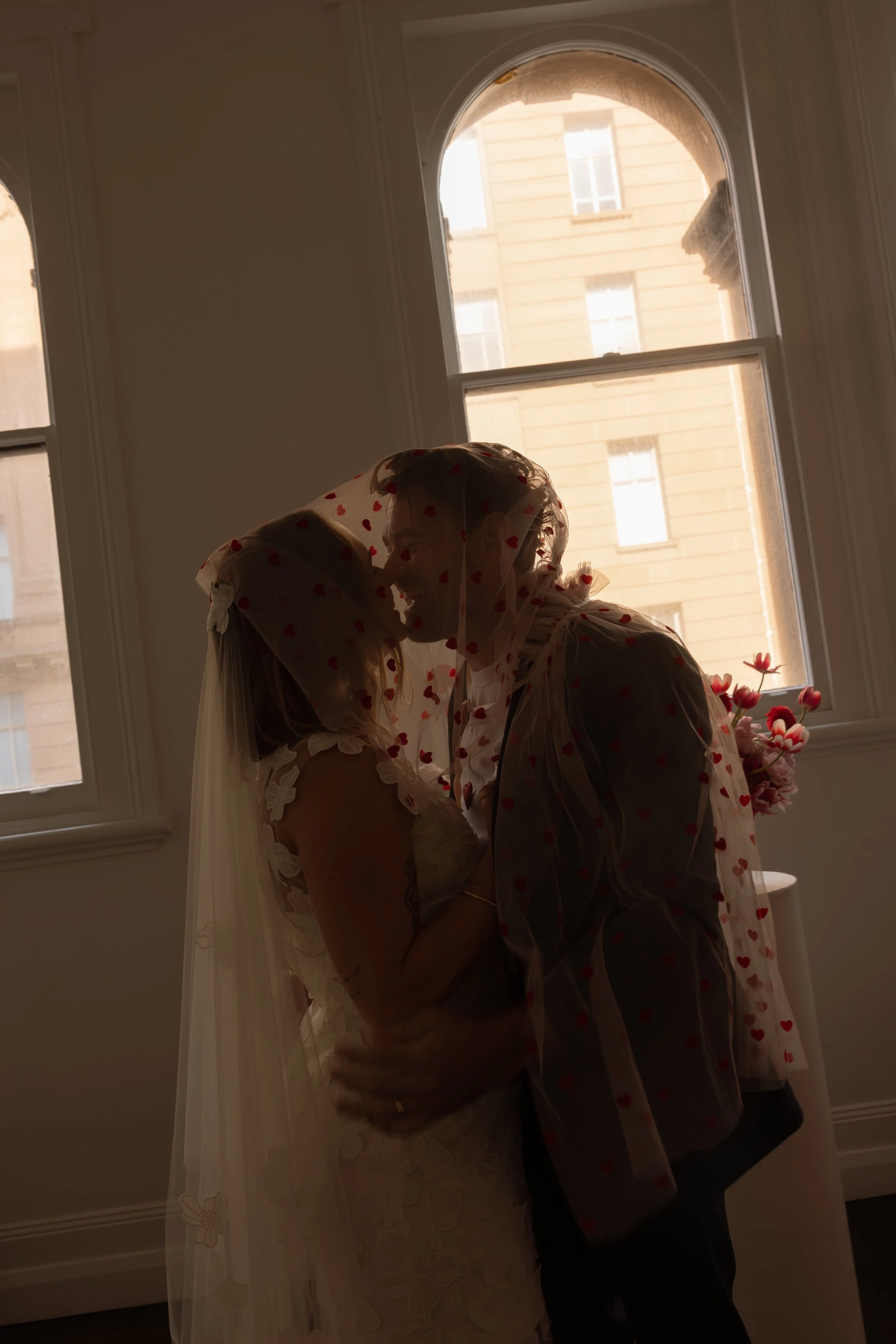 Silhouette of a couple kissing by a window, with a bride holding flowers and wearing a veil with red hearts, backlit by natural light.