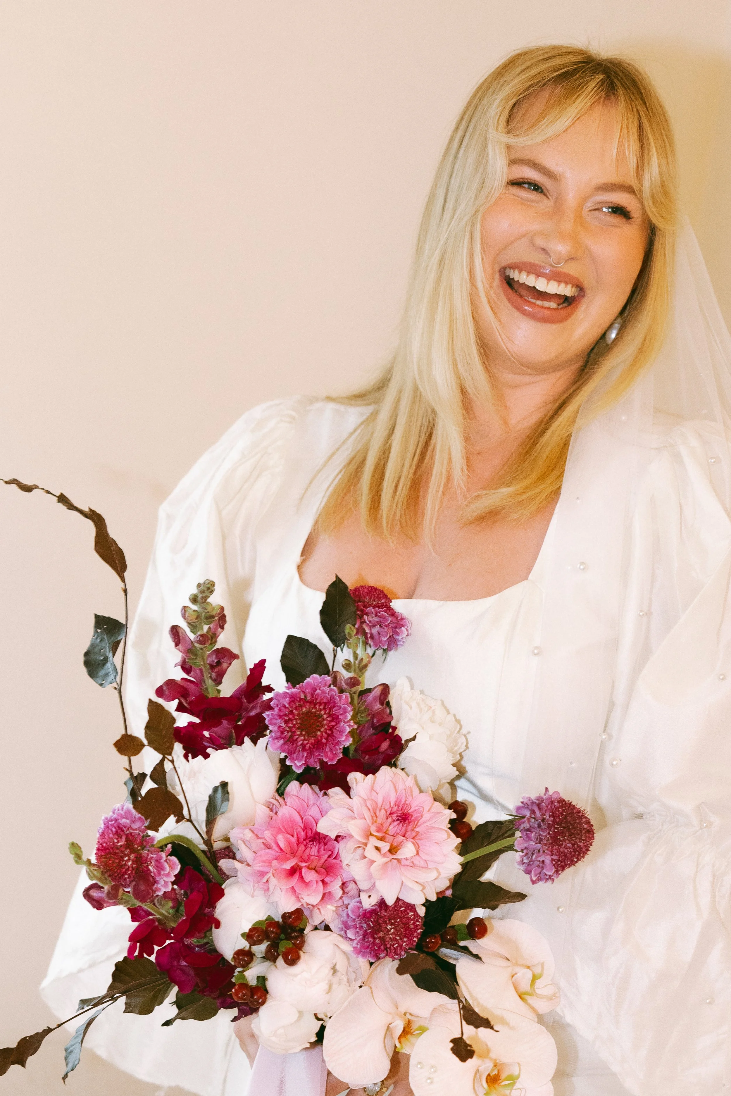 A woman with blonde hair and a wide smile, wearing a white dress and holding a bouquet of pink, white, and purple flowers.