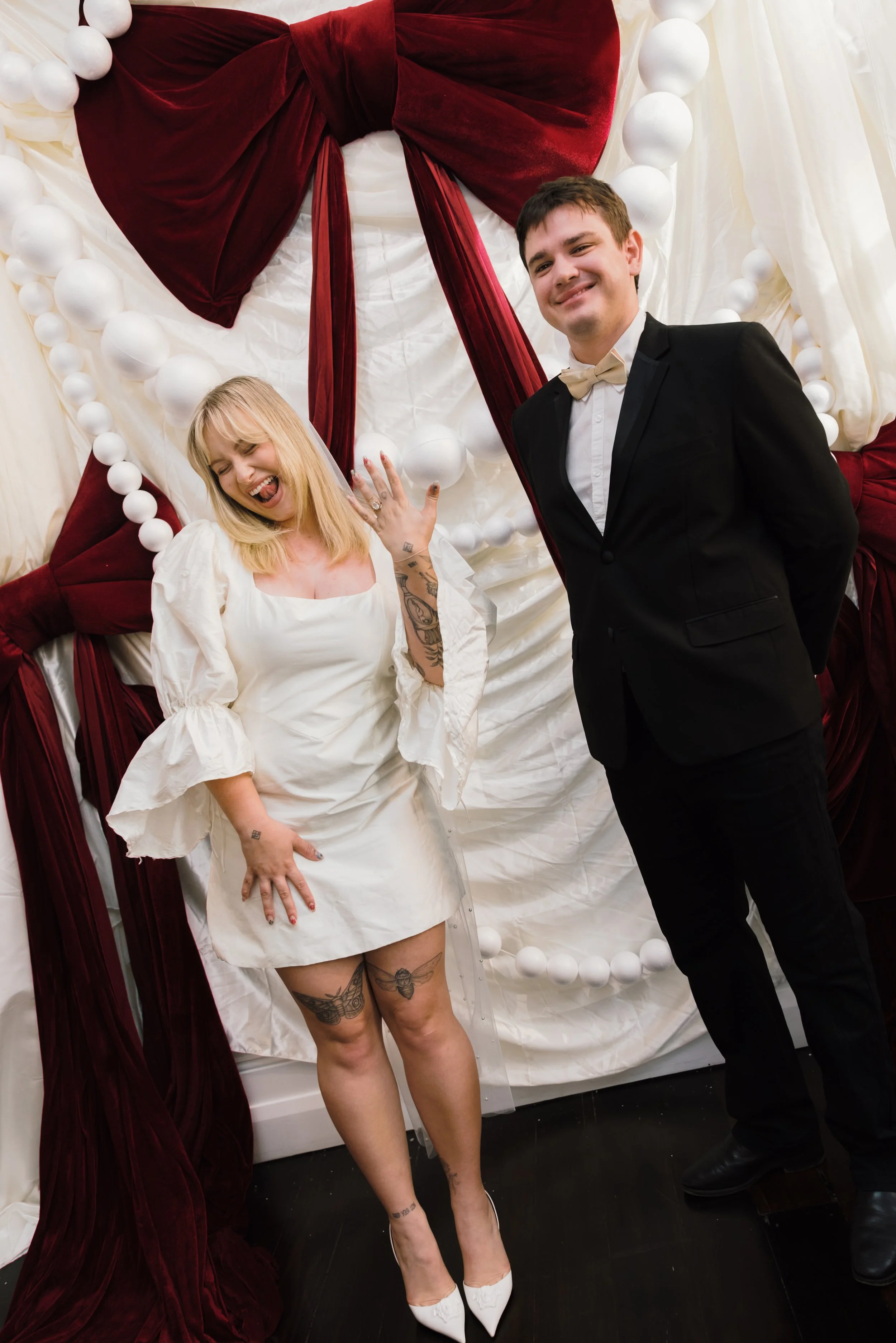 Two people posing in front of decorated backdrop with white fabric, large red bows, and white ball garlands. The woman has blonde hair, tattoos, and is wearing a white dress with puffed sleeves, showing her tattoos. She is laughing with one hand on h