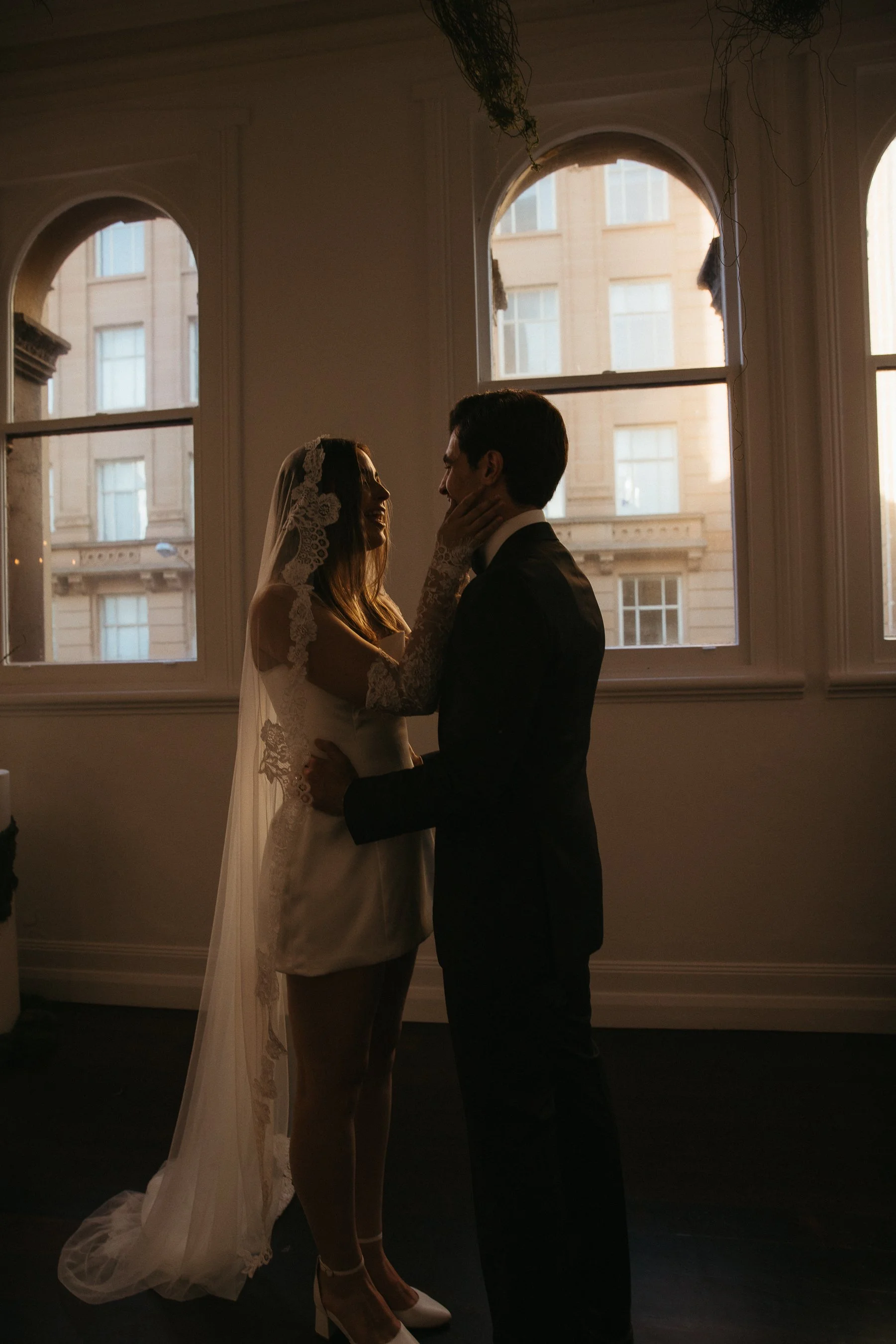 A bride and groom holding each other, smiling, standing in front of windows with sunlight shining through during their wedding.