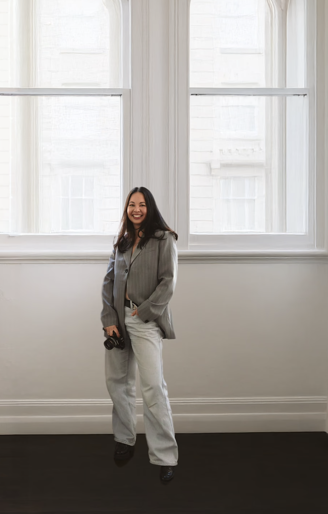 Woman smiling and holding a camera, standing in front of large window with white curtains in a bright room.