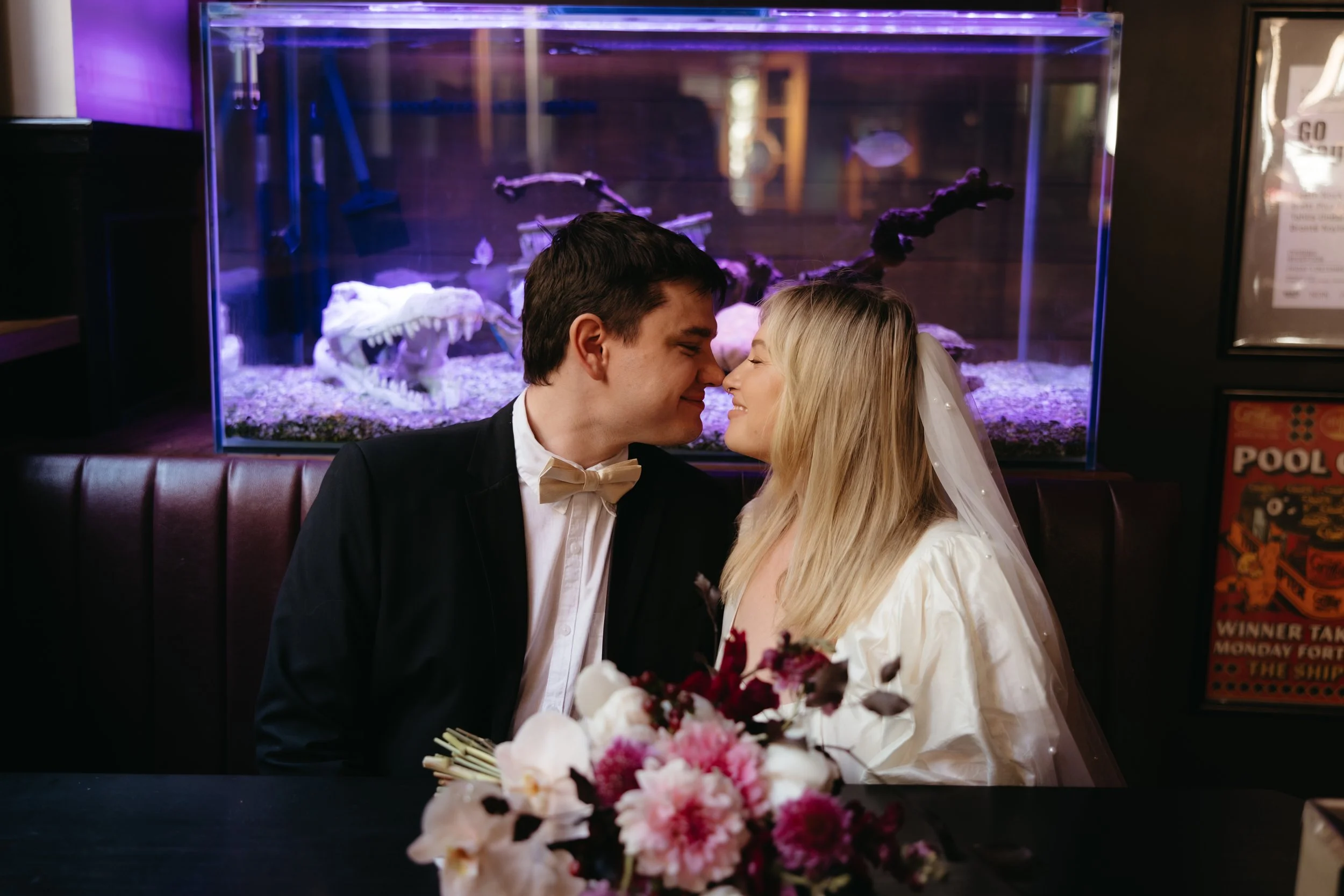 A couple dressed in wedding attire, leaning close and touching noses, sitting at a table with a flower bouquet in front of them in front of an aquarium with white crocodile and fish, in a dimly lit restaurant.