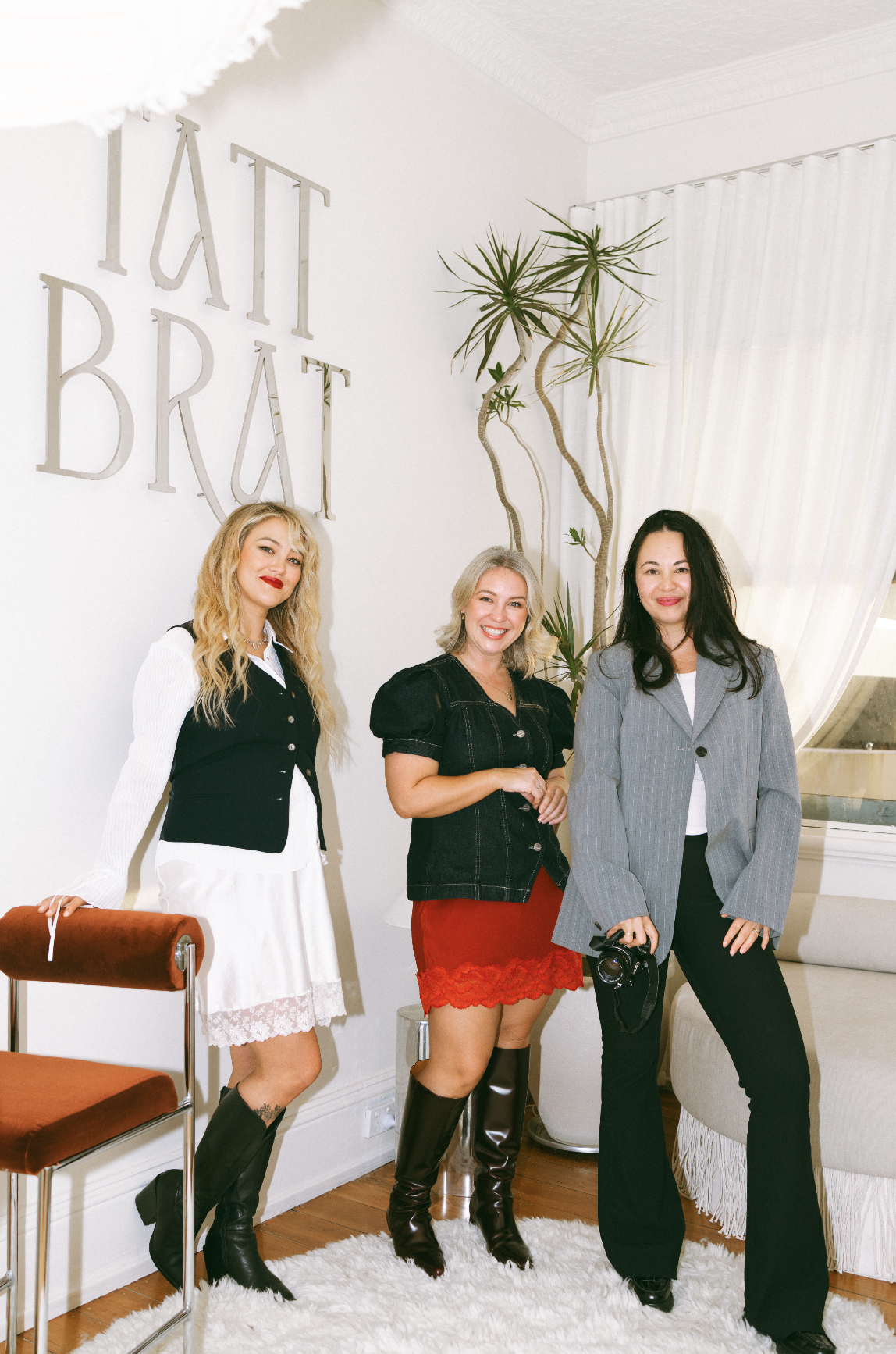 Three women standing indoors, smiling, with a wall sign that says 'TATT BRÁT' and a large potted plant nearby.