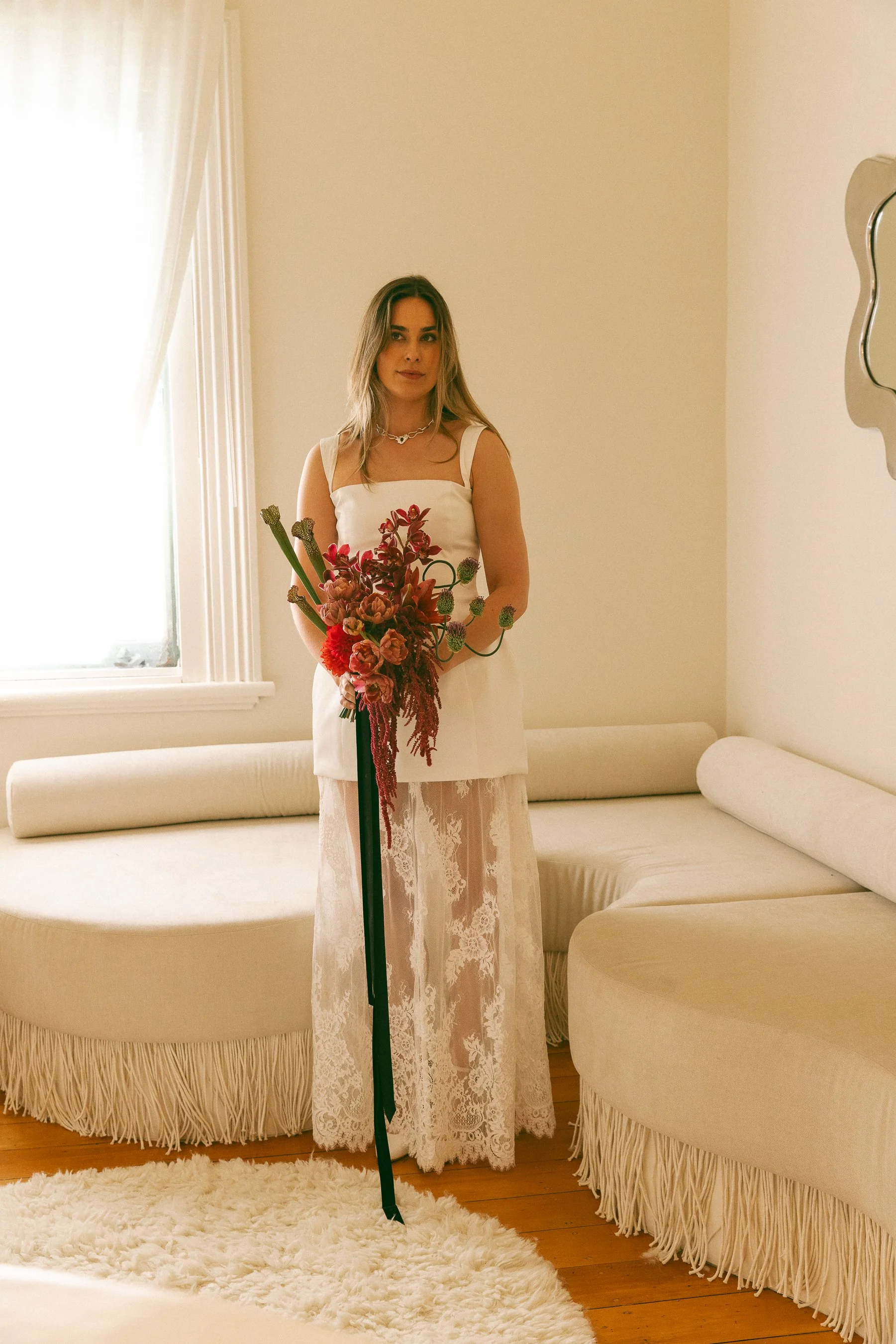A woman in a white dress with lace details, holding a bouquet of red flowers, standing in a warmly lit room with cream-colored walls and furniture.