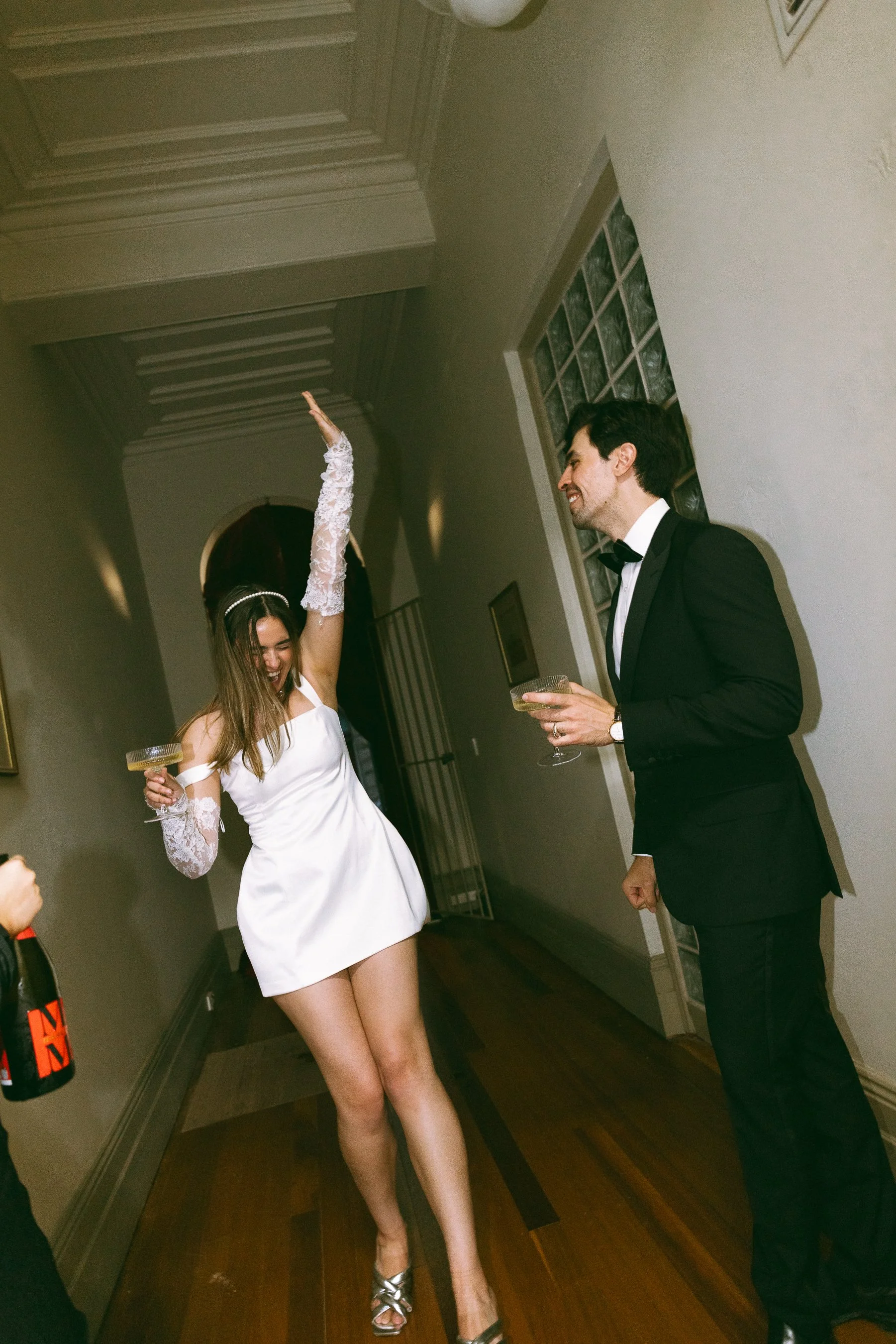 Man and woman in formal attire celebrating at a party, woman in a white dress with lace gloves raising her arm, man in a black tuxedo holding a glass, in an elegant hallway.