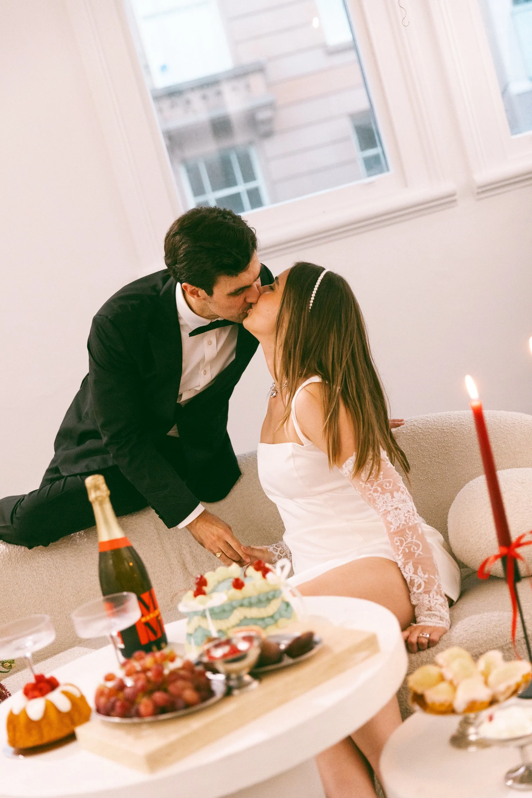 A man in a tuxedo and a woman in a white dress kissing on a sofa during a celebration, with a table of desserts, a cake, and a bottle of champagne nearby.