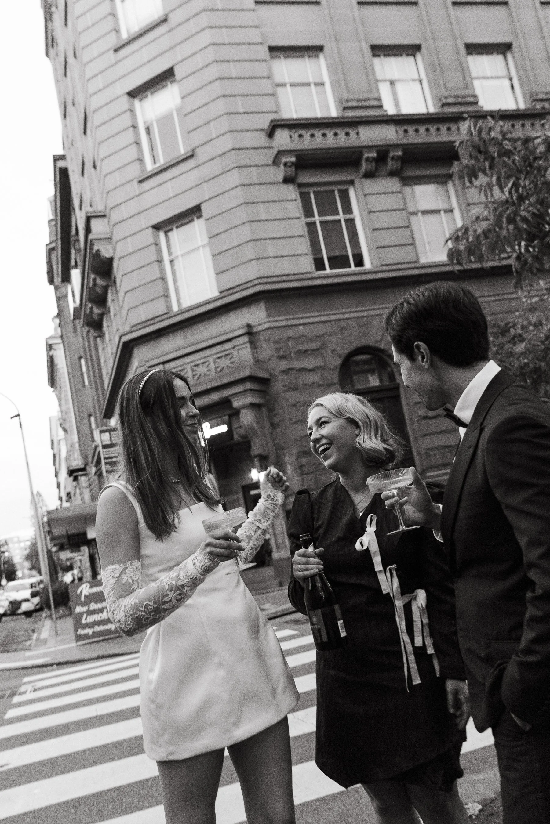 Three people in formal attire having drinks and talking on a city street at night, with a tall building in the background.
