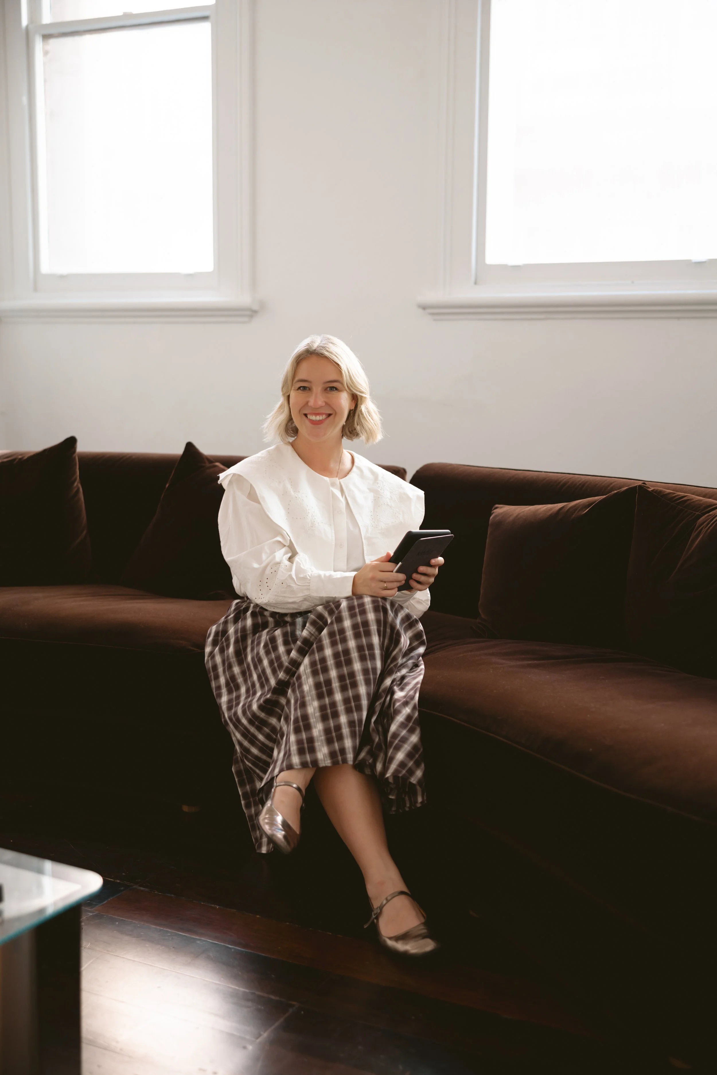A woman with blonde hair smiling and sitting on a dark brown couch, holding a smartphone, in a bright room with white walls and two large windows.