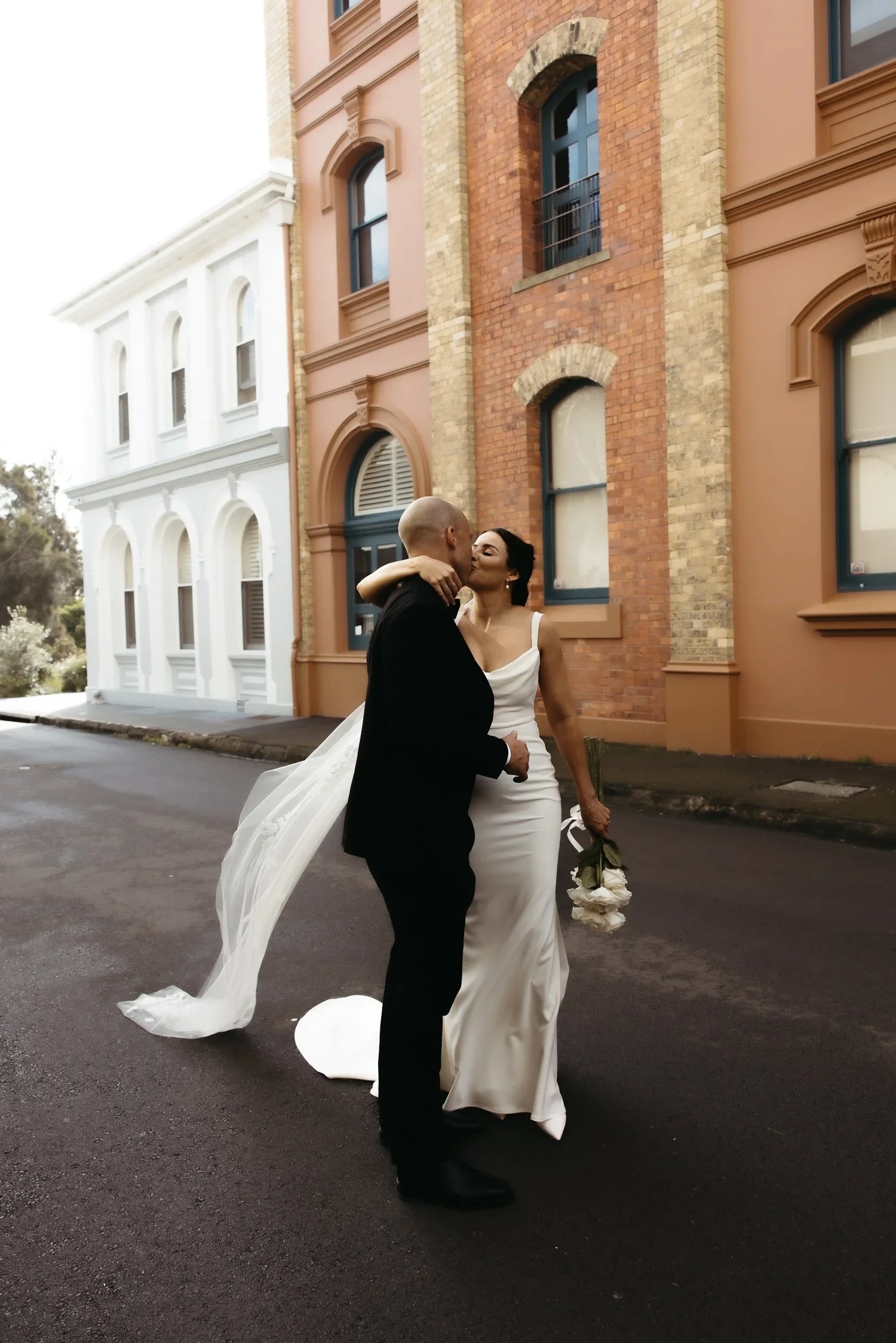A newlywed couple sharing a kiss on a city street, with the groom in a black suit and the bride in a white wedding dress holding a bouquet, in front of colorful brick and white buildings.
