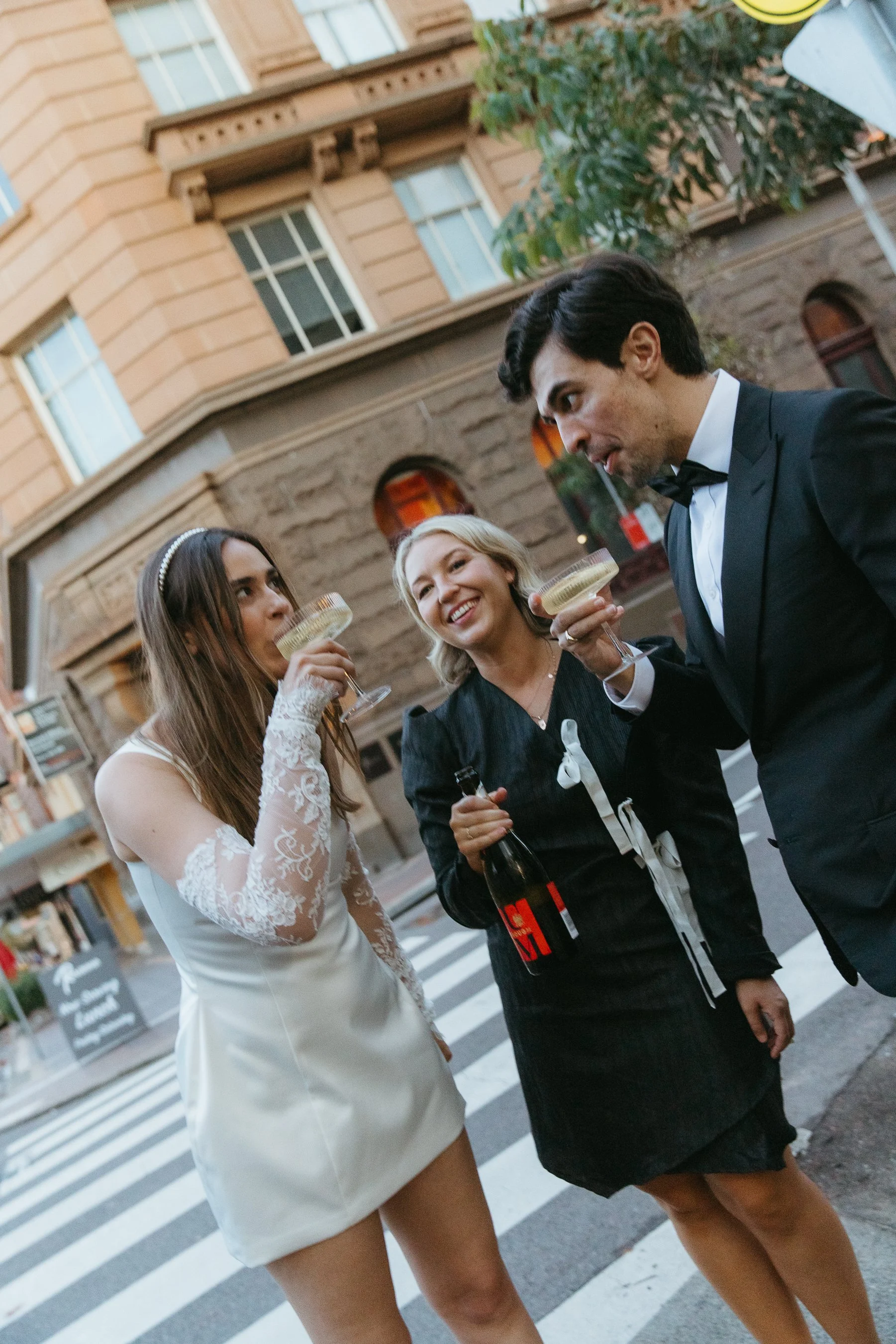 Three people in formal attire enjoying drinks together on a city street, with a building and trees in the background.