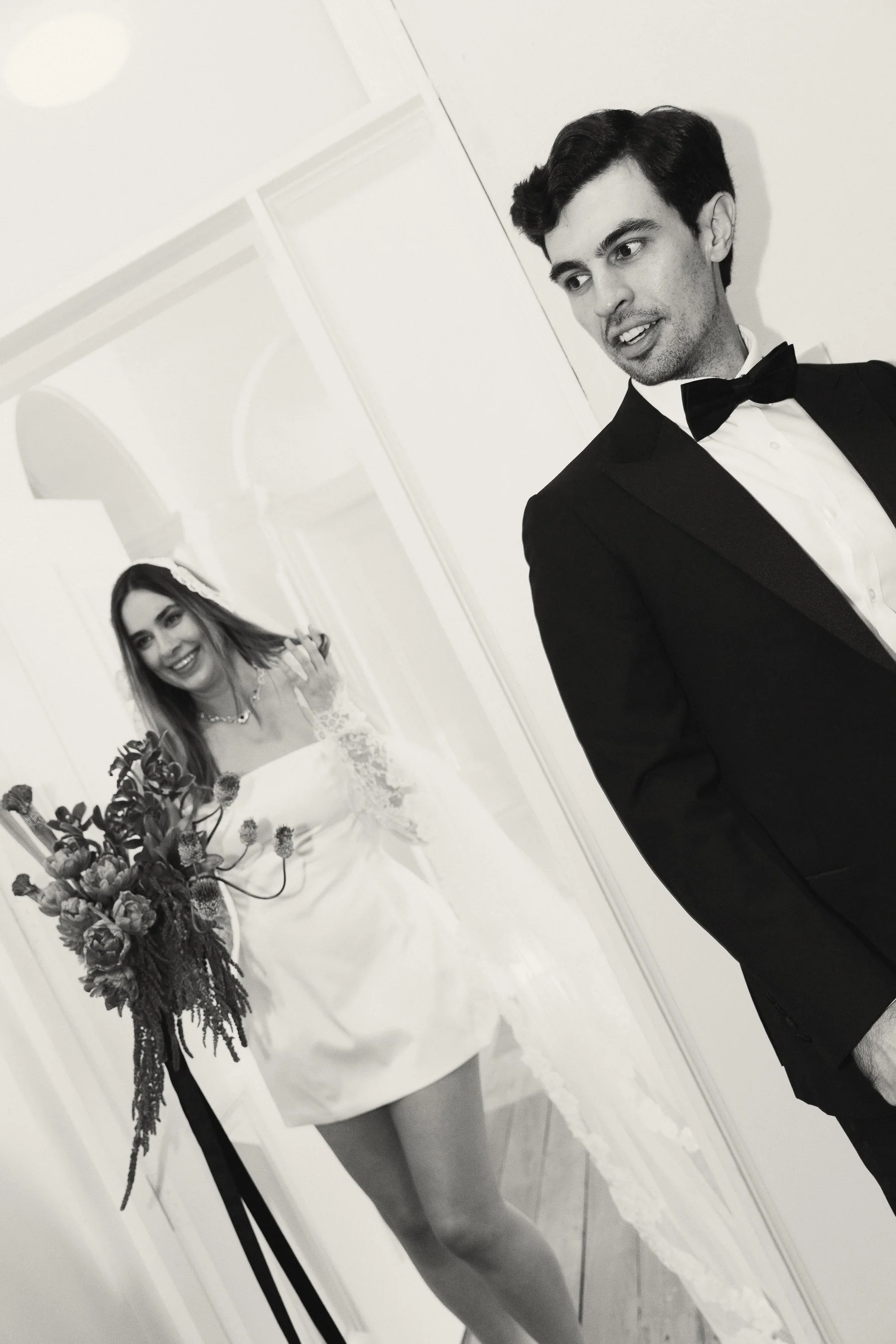 A mirror reflection of a woman in a wedding dress holding a bouquet and a man in a tuxedo getting ready for a wedding.