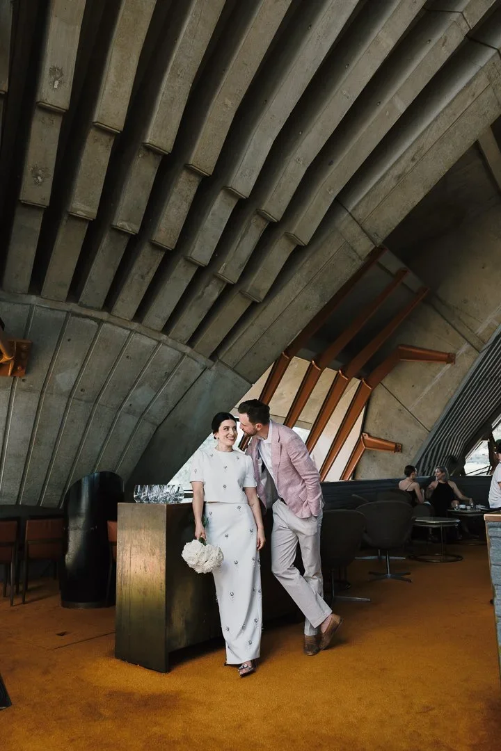 A couple in formal attire, with the woman holding a bouquet of white flowers, stands inside a modern building with concrete and wood architectural features, and other people seated in the background.
