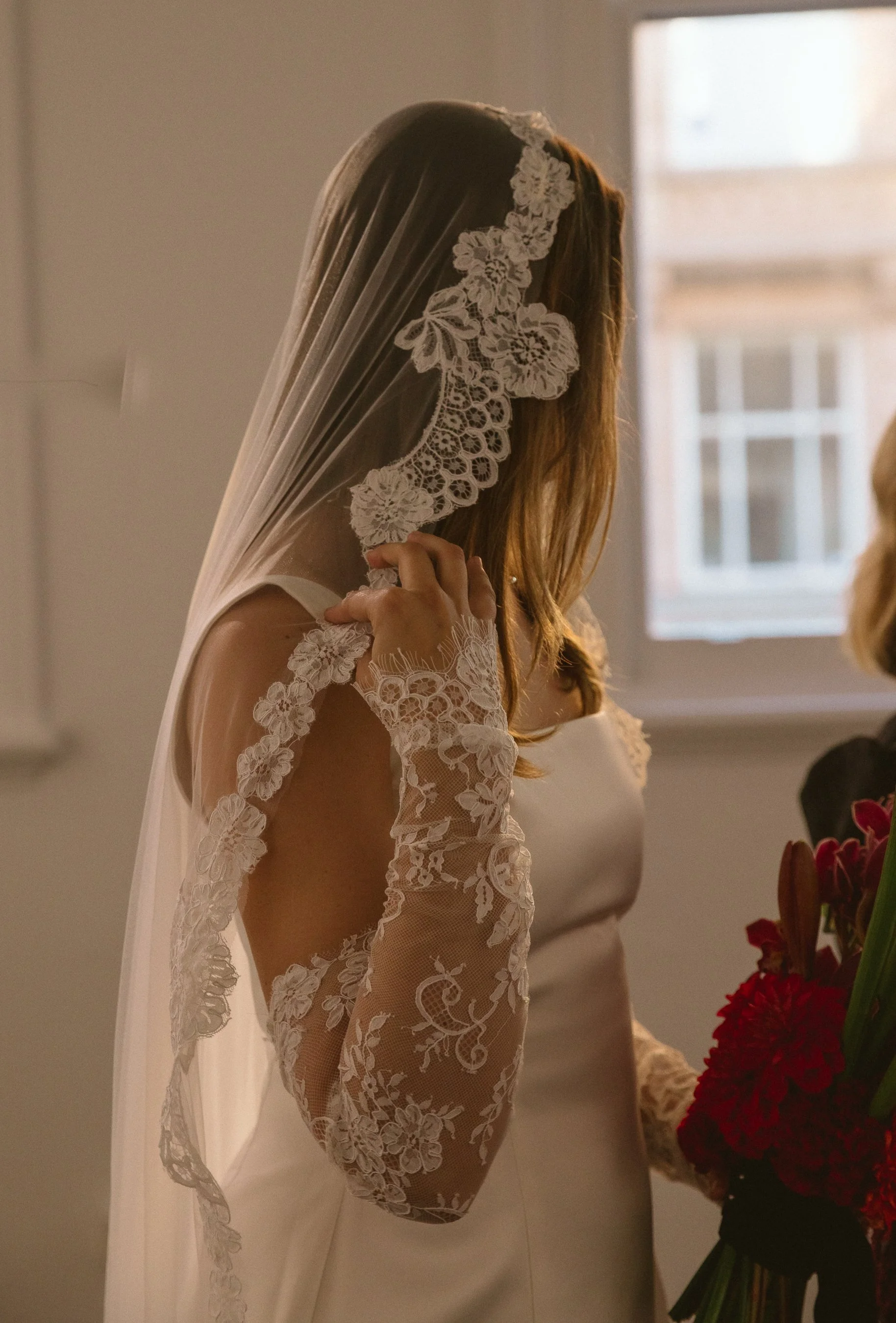 A bride wearing a lace wedding gown and a veil, holding a bouquet of red flowers, indoors near a window.