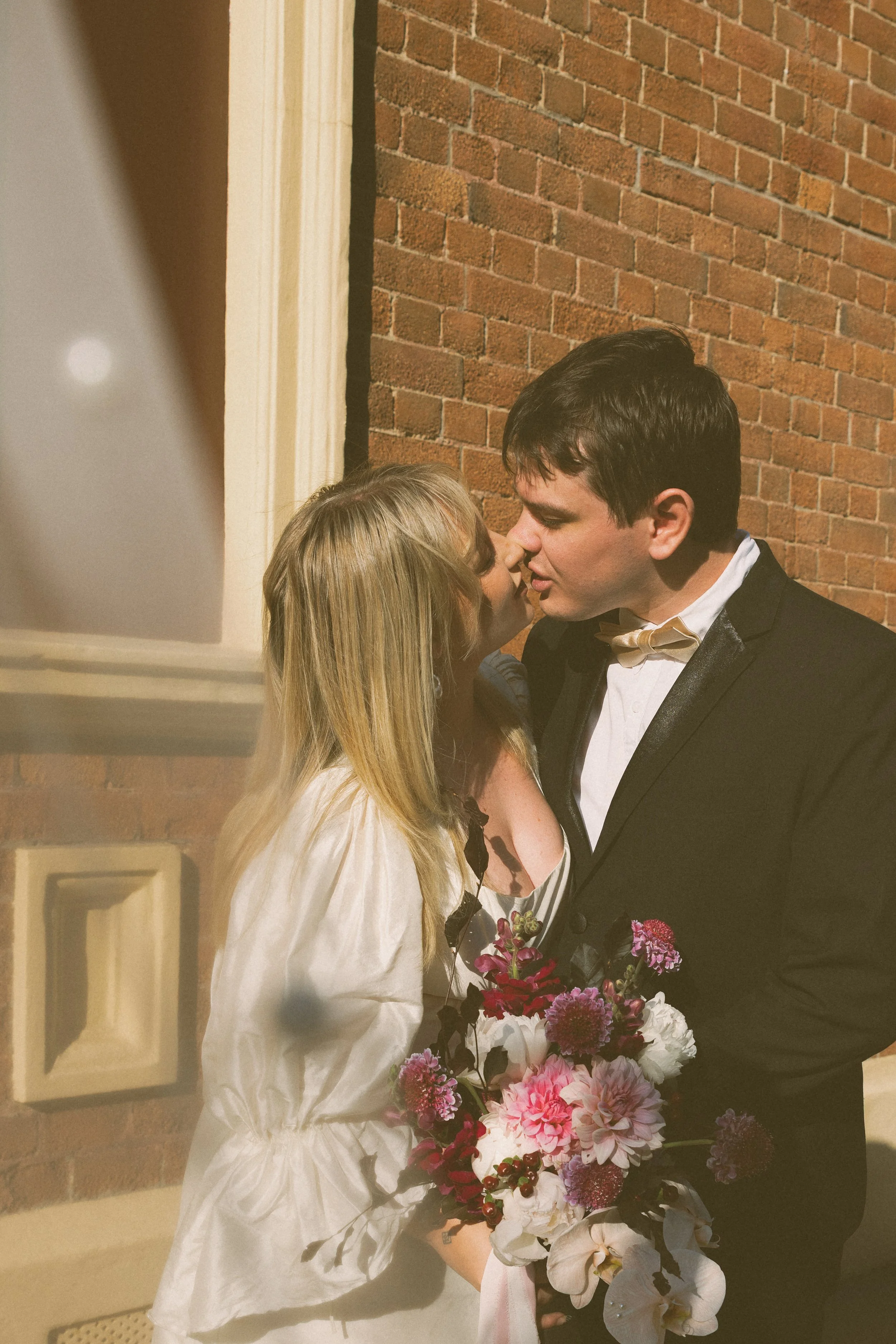 A man and a woman are about to kiss outside a brick building, with the woman holding a bouquet of pink, white, and purple flowers.