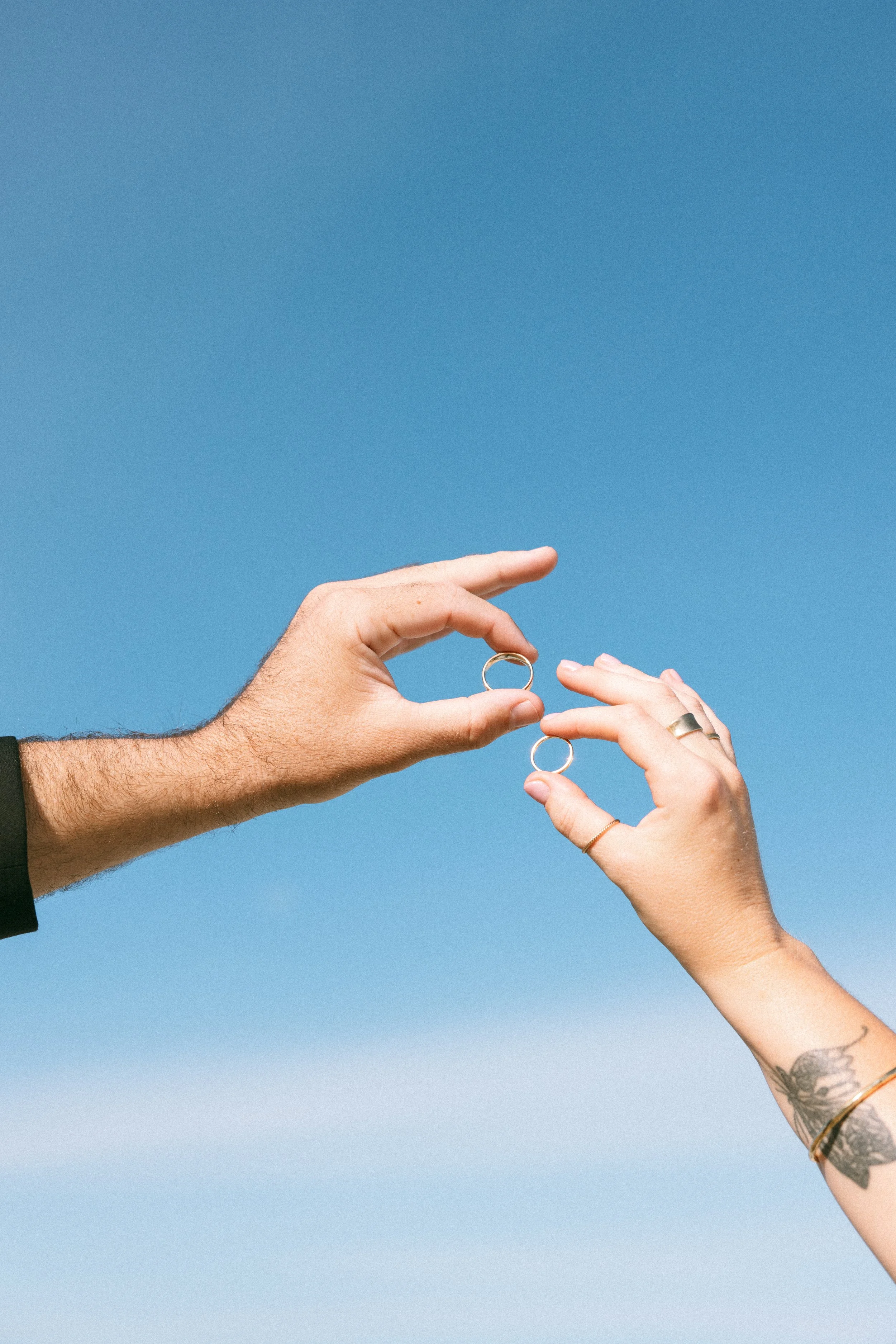 Two hands exchanging wedding rings against a clear blue sky, one hand has a tattoo on the wrist and wears a ring, the other hand has a ring on the finger.