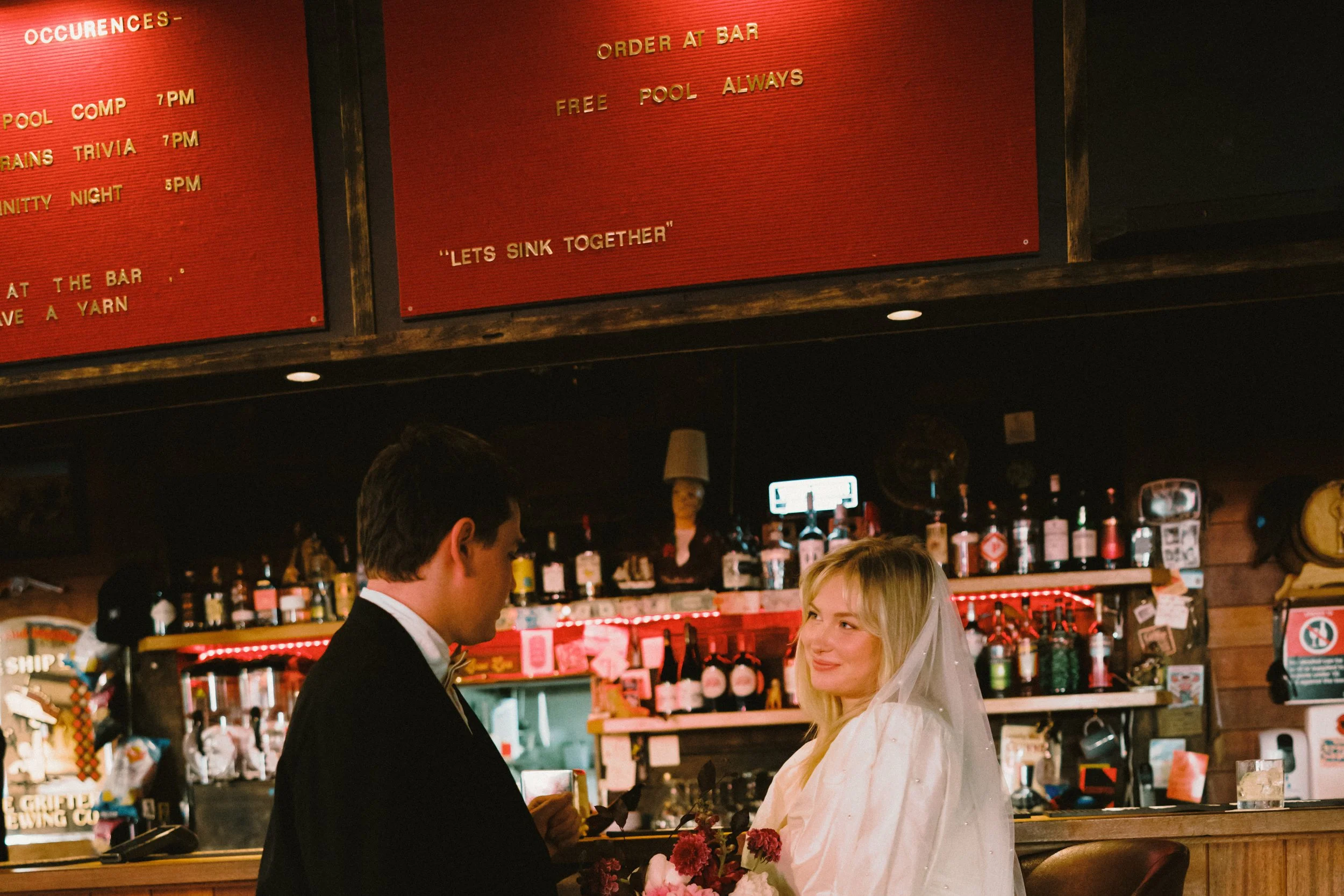 A man and woman in formal attire sharing a moment at a bar, with bottles of alcohol on shelves behind them and a red menu board above.