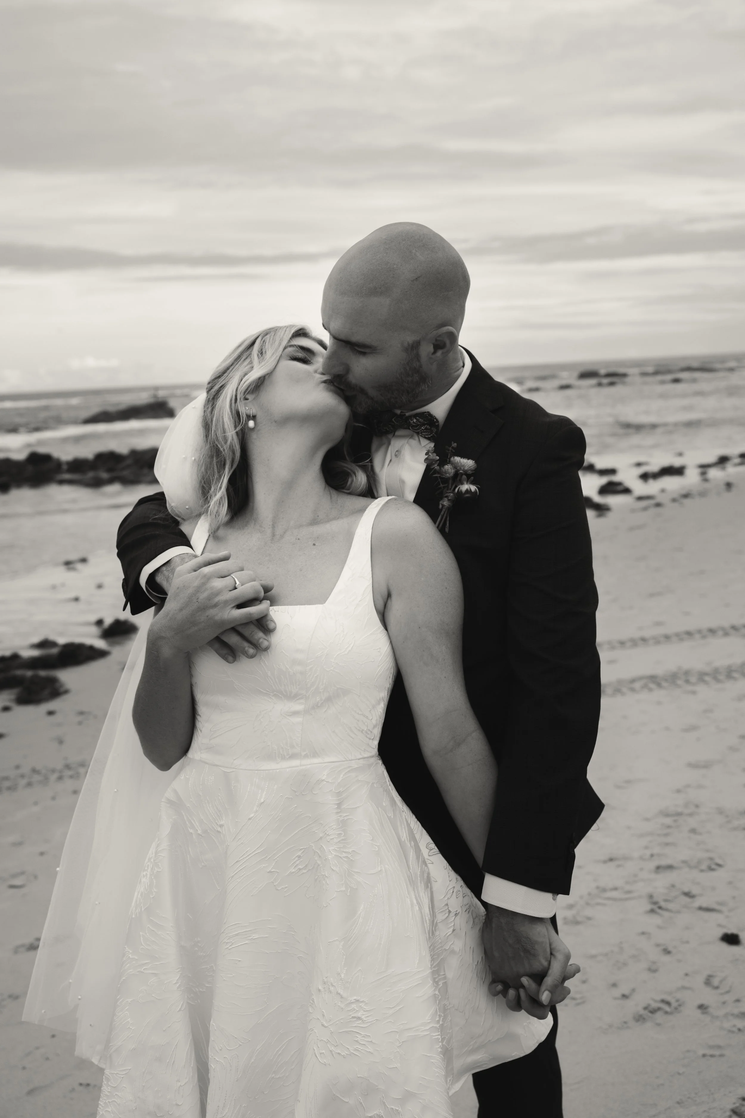 A couple in wedding attire sharing a kiss on a beach, with the ocean and a cloudy sky in the background.