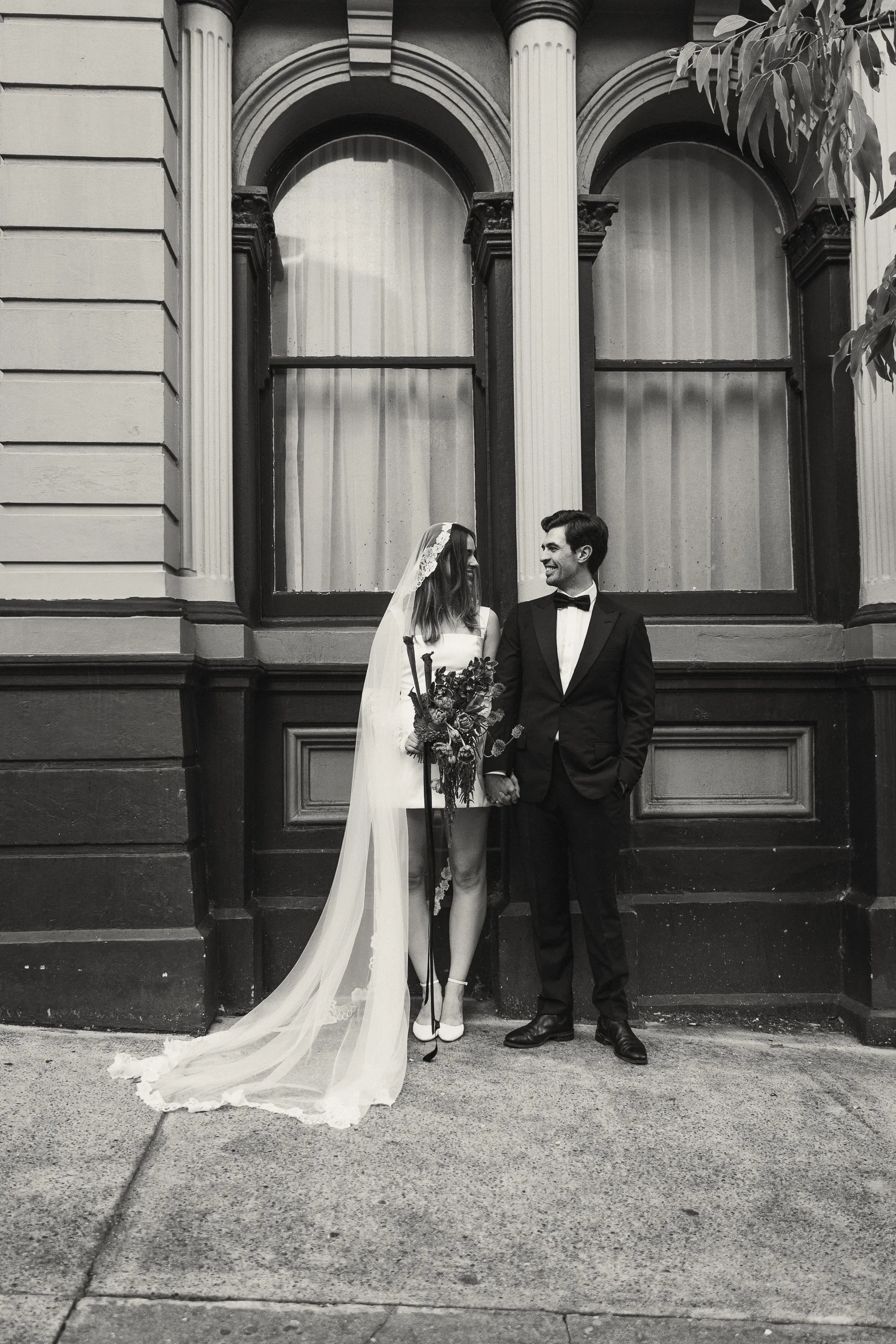 A bride and groom standing against a building with large arched windows, holding hands and smiling at each other in black and white.