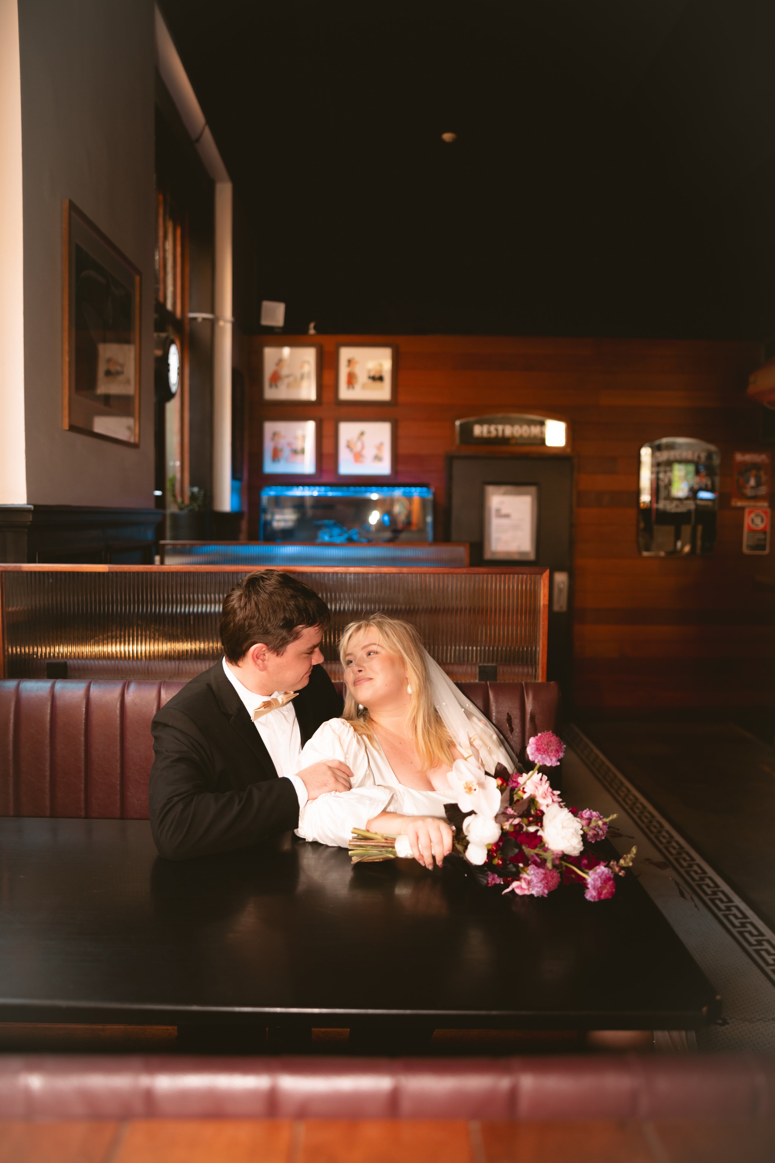 A bride and groom sitting closely at a restaurant table, gazing into each other's eyes, with a bouquet of flowers in front of them.