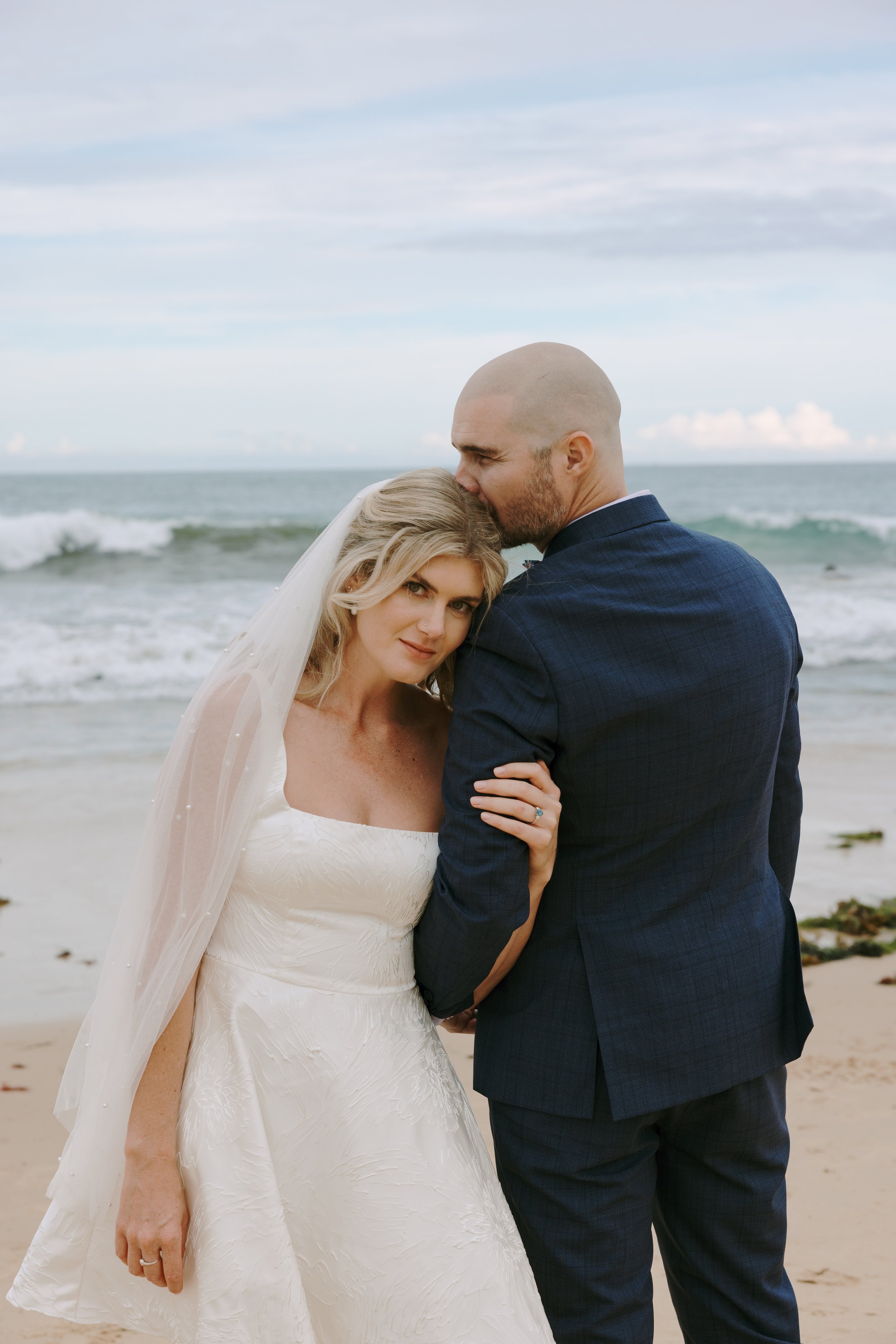 A bride and groom on a beach, with the groom kissing the bride's forehead, the bride wearing a white wedding dress and veil, and the groom in a dark suit near the ocean.