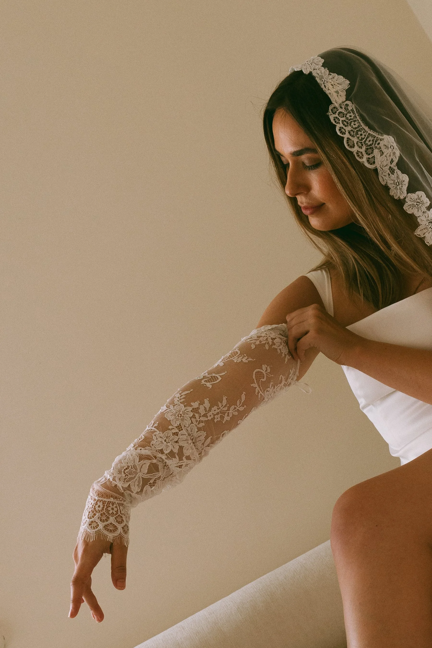 A bride in a wedding dress and veil, adjusting her lace-sleeved arm in a soft, indoor setting.