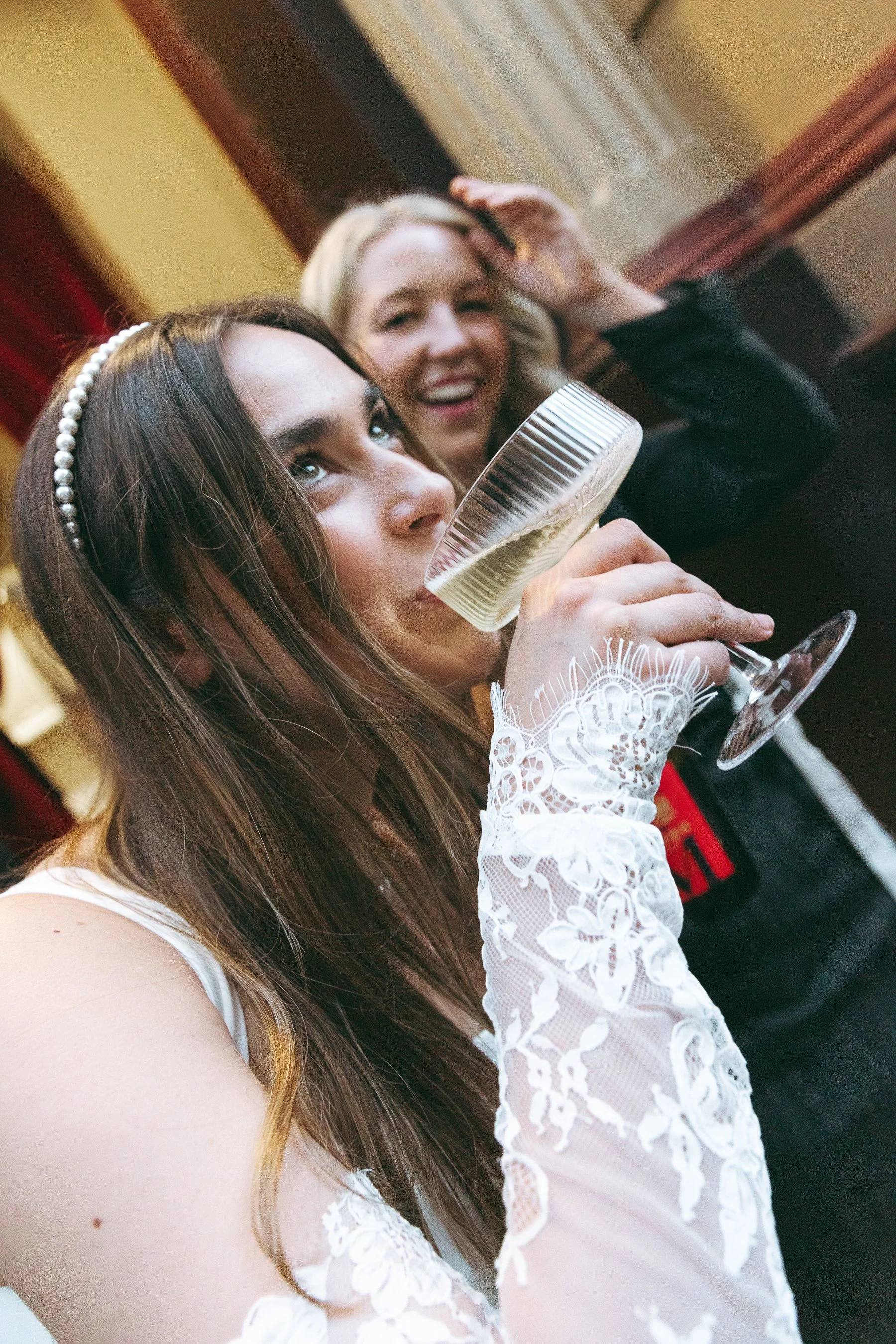 A young woman with long brown hair and a pearl headband drinks champagne from a flute at a lively gathering, with another woman smiling and waving in the background.