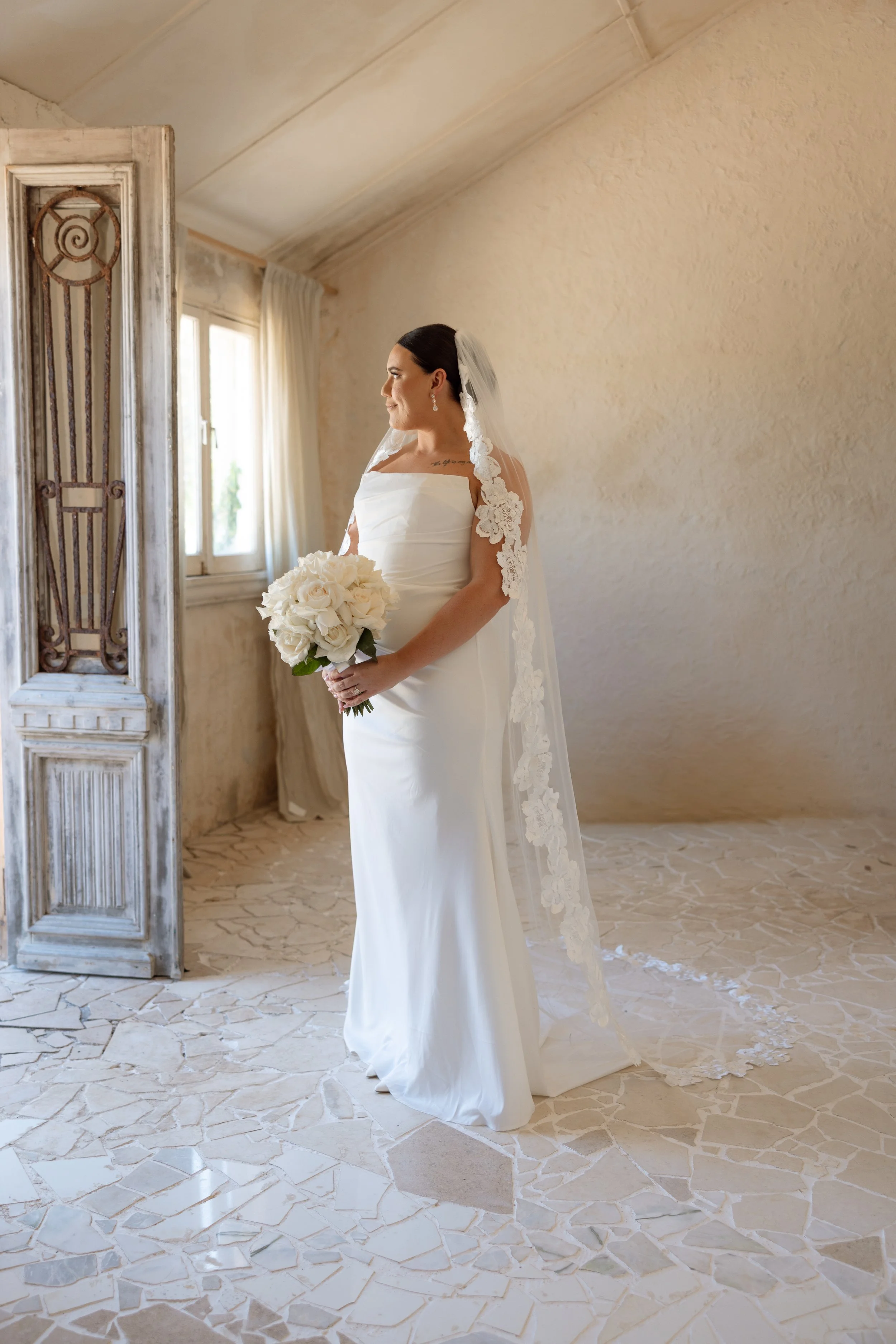 A bride in a white wedding dress holding a bouquet of white roses, standing in a sunlit room with beige textured walls and a rustic wooden door.