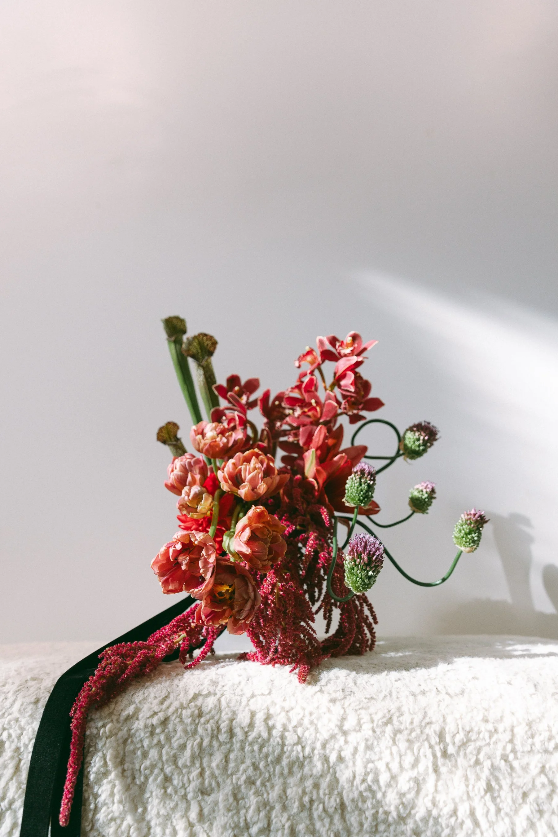 A bouquet of pink, red, and purple flowers resting on a soft, white textured surface with a plain light-colored wall in the background.