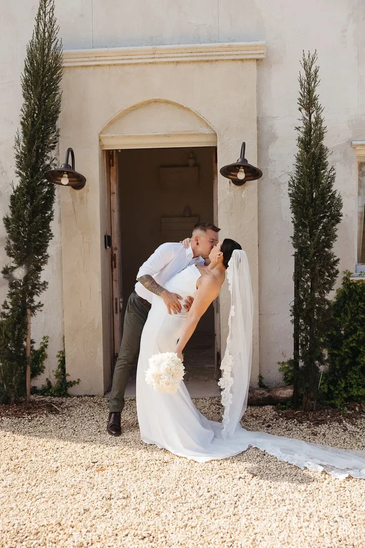 A newlywed couple shares a kiss outside a building with two trees and vintage wall lighting.