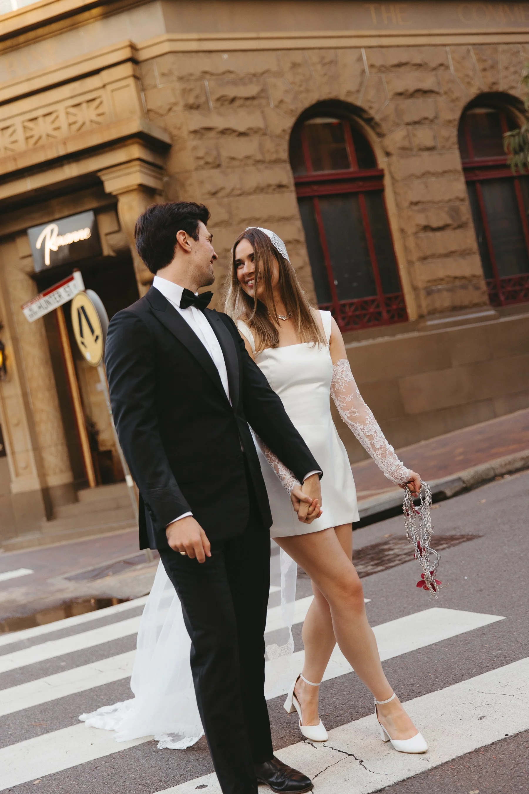 Couple dressed in wedding attire holding hands and walking across a pedestrian crosswalk on a city street, smiling at each other.