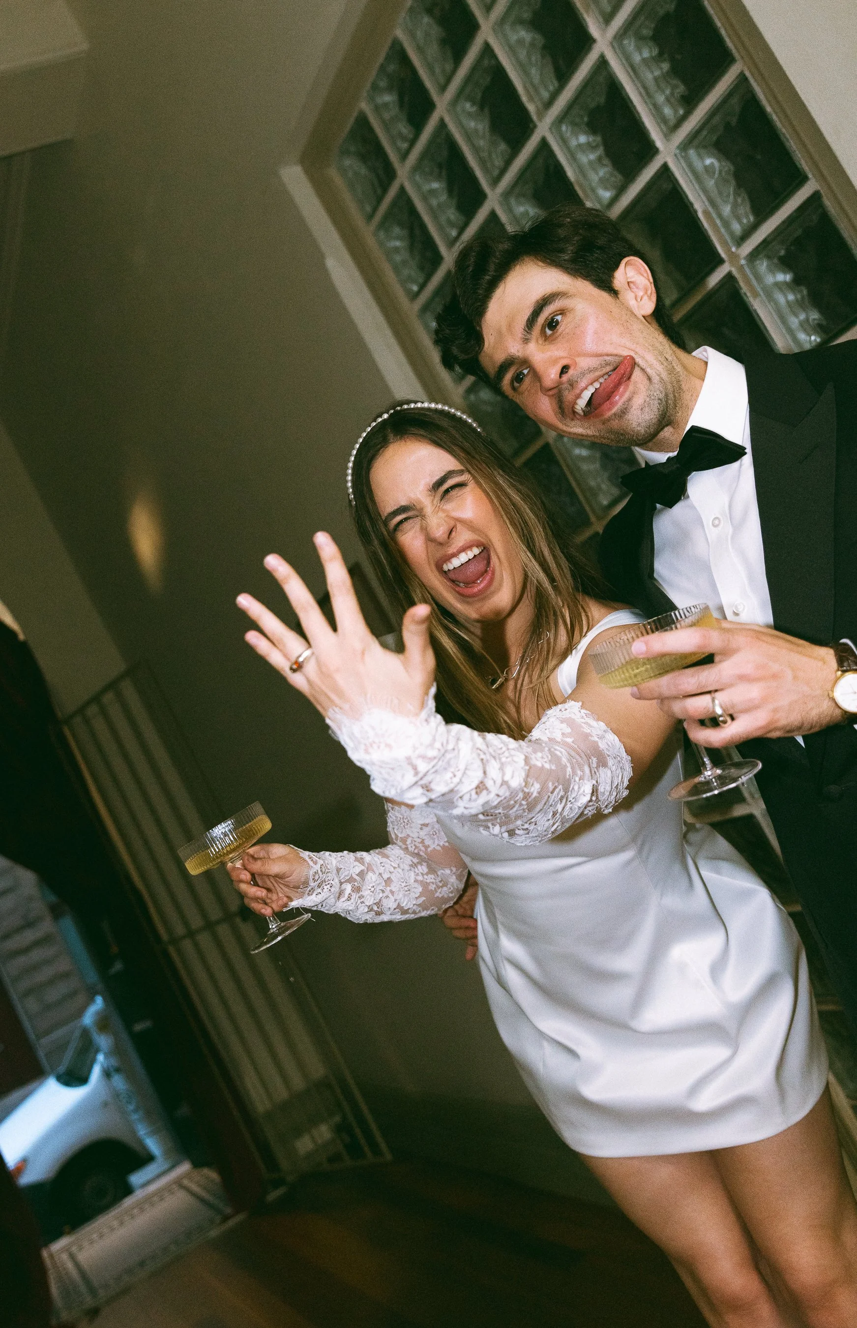 A couple in wedding attire celebrating at a party, holding drinks, with playful expressions and making silly faces.
