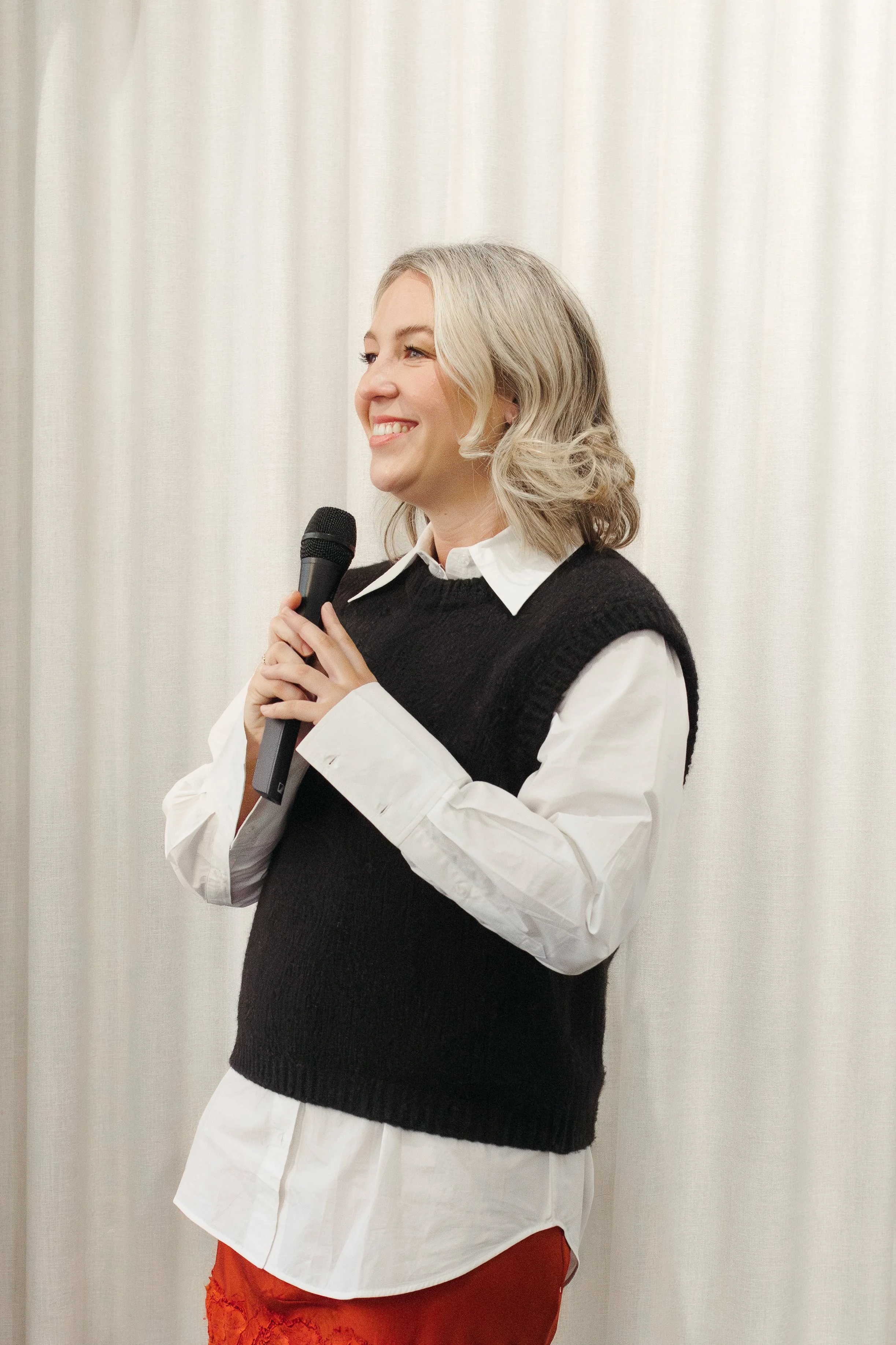 Woman holding microphone and smiling at an event in front of white curtain.