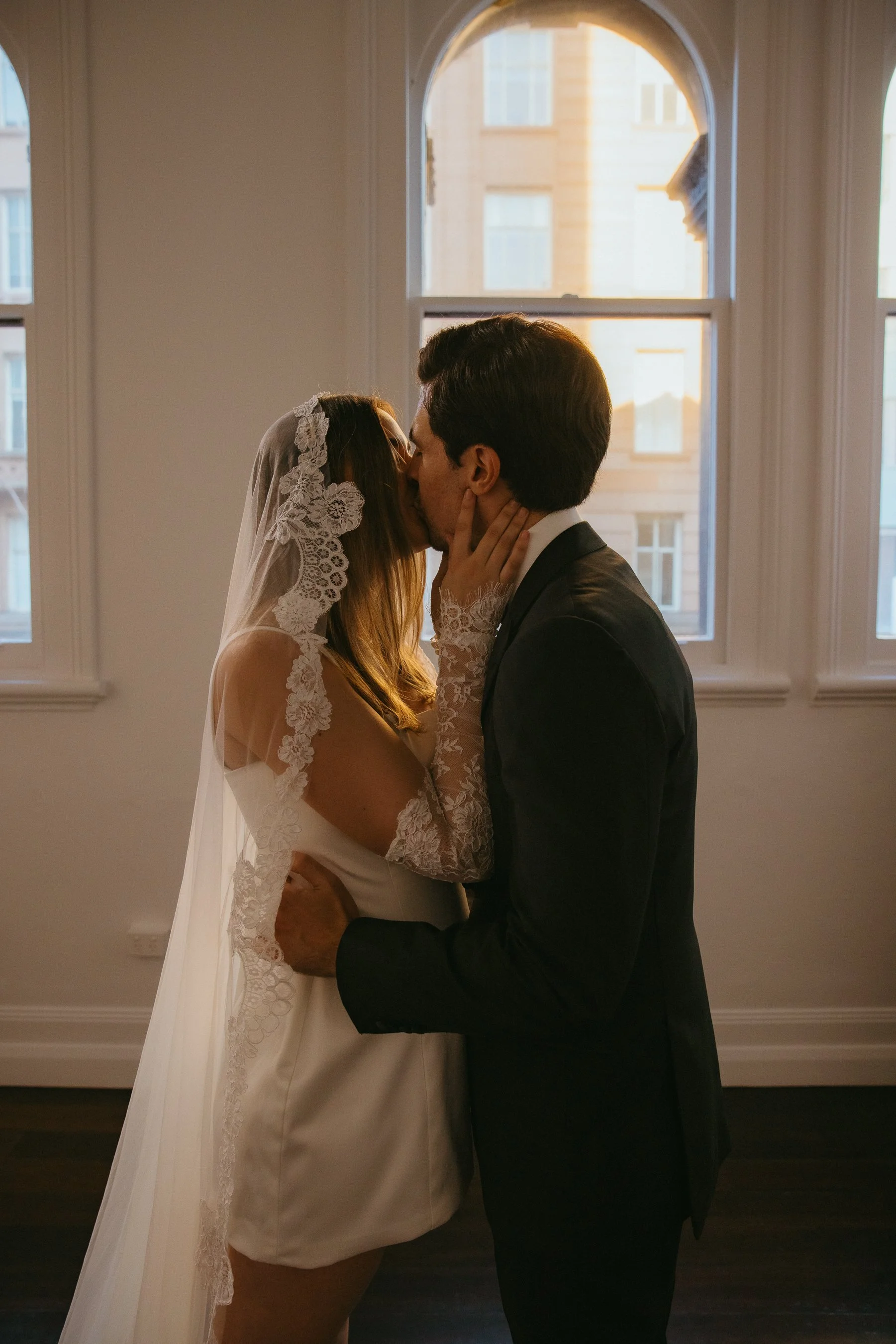 A bride and groom sharing a kiss indoors, backlit by a large window, with the bride wearing a lace veil and dress and the groom in a dark suit.