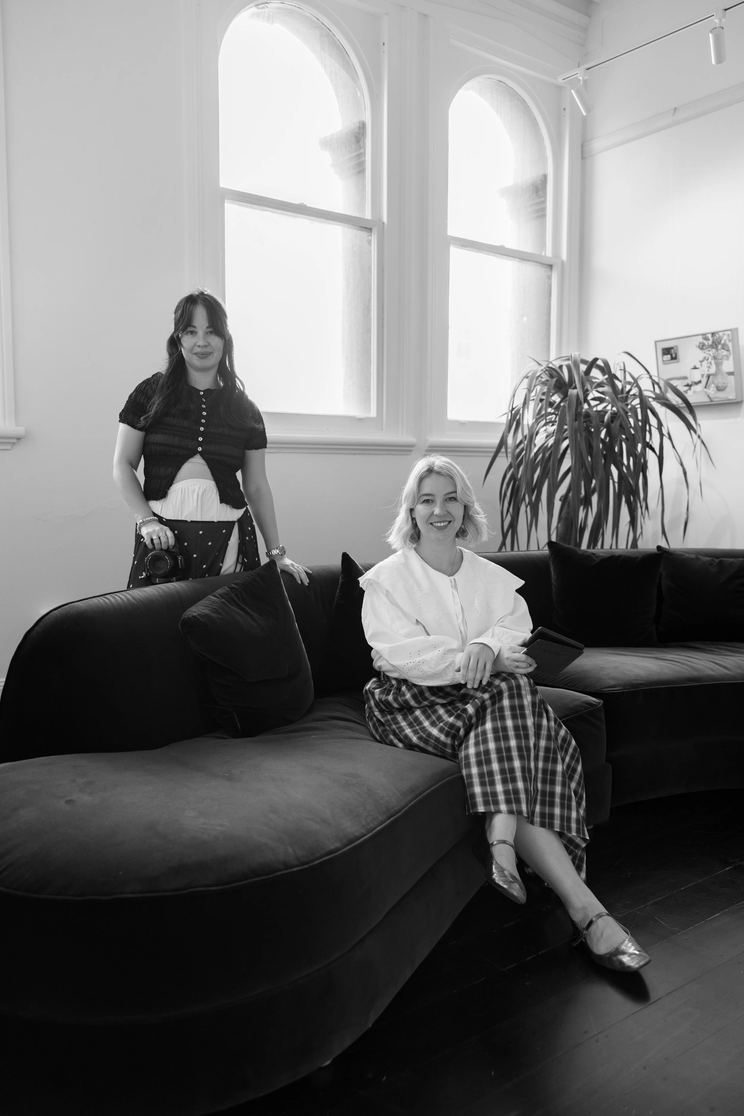 Two women in a living room with high arched windows, a large potted plant, and artwork on the wall. One woman is sitting on a couch holding a smartphone, while the other stands beside her with a camera hanging from her neck.