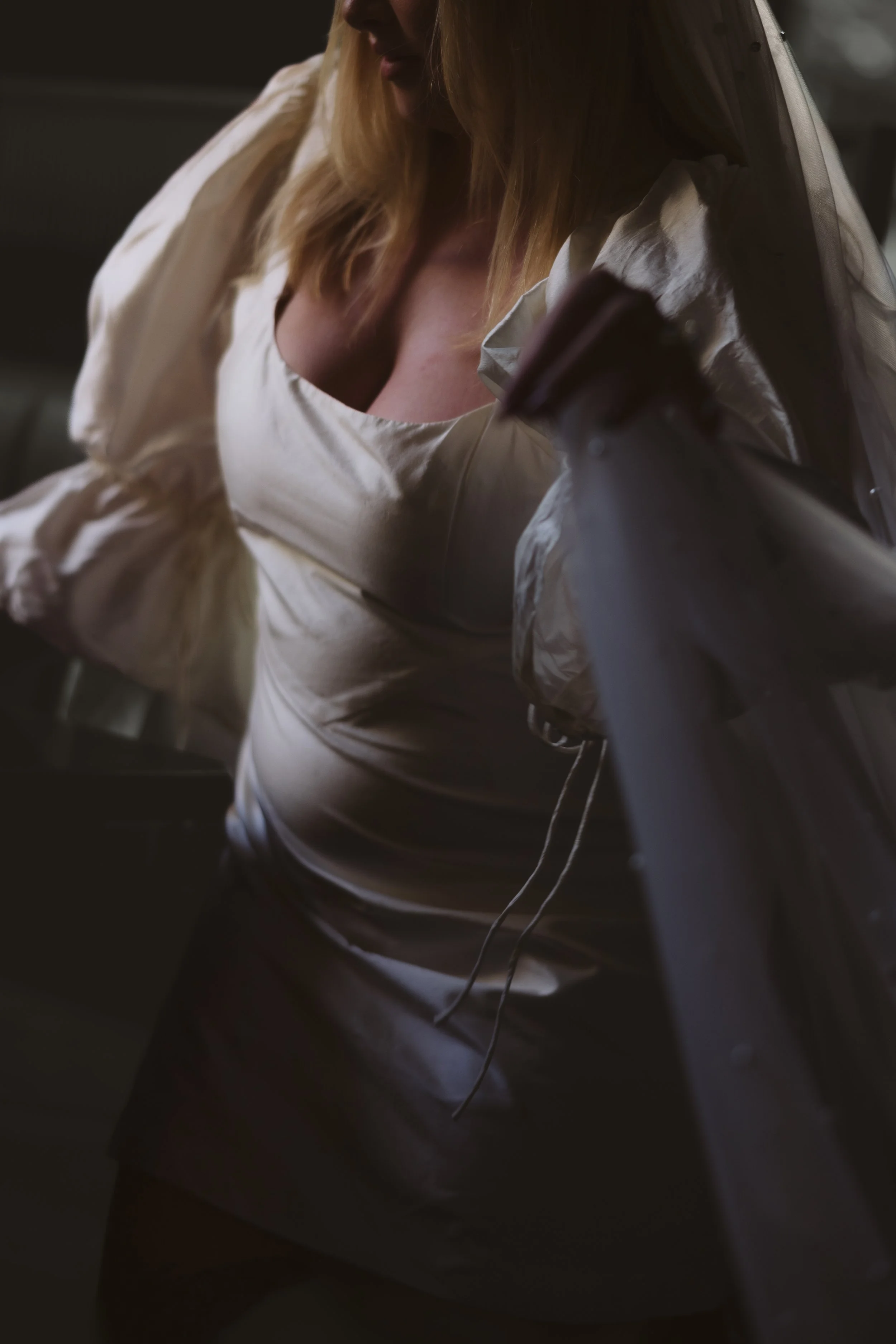 A woman with blonde hair wearing a satin cream-colored top, standing in dim lighting with a dark background, partially obscured by a dark object in the foreground.