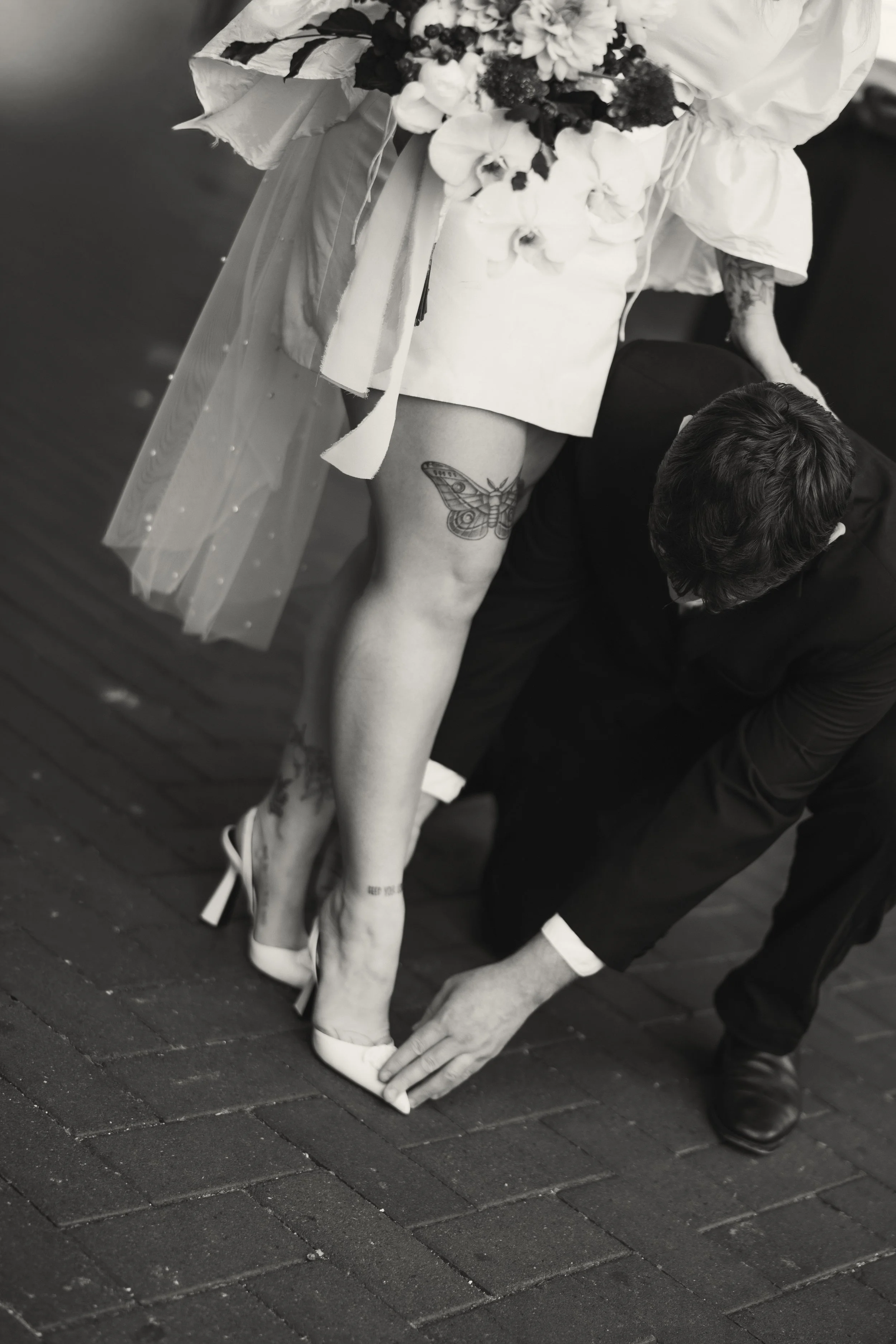A bride wearing high heels and a floral dress getting her shoes fitted by a man dressed in a suit. The bride has a butterfly tattoo on her upper thigh and other tattoos on her calves.