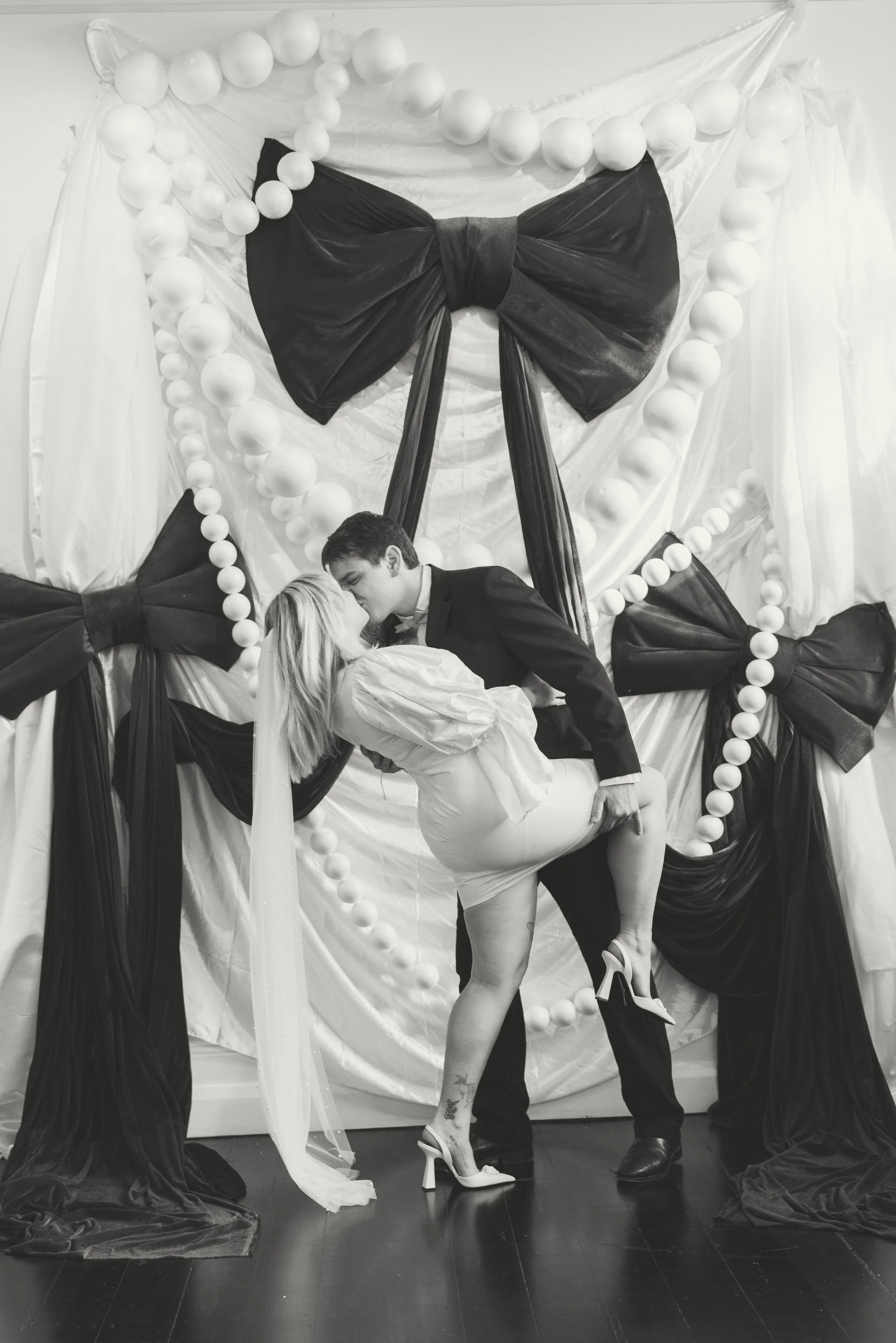 A black and white photo of a man in a tuxedo kissing a woman in a light-colored dress who is bent backward with her legs wrapped around his waist. They are in front of a decorated backdrop with large black bows and string lights.