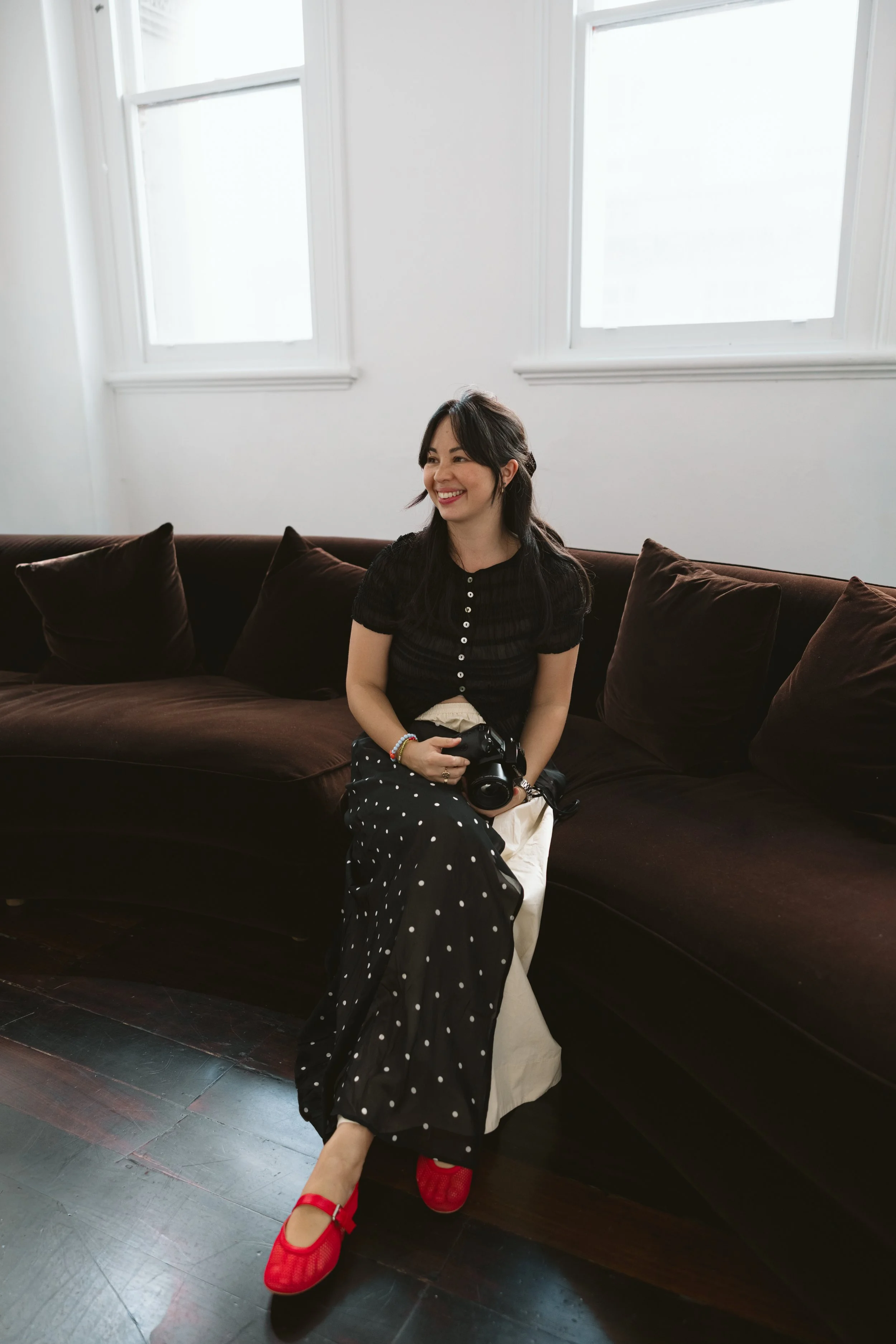 A woman with dark hair, wearing a black top with buttons and a black skirt with white polka dots, sitting on a dark brown couch in a room with white walls and windows, holding a camera, smiling, and wearing red shoes.
