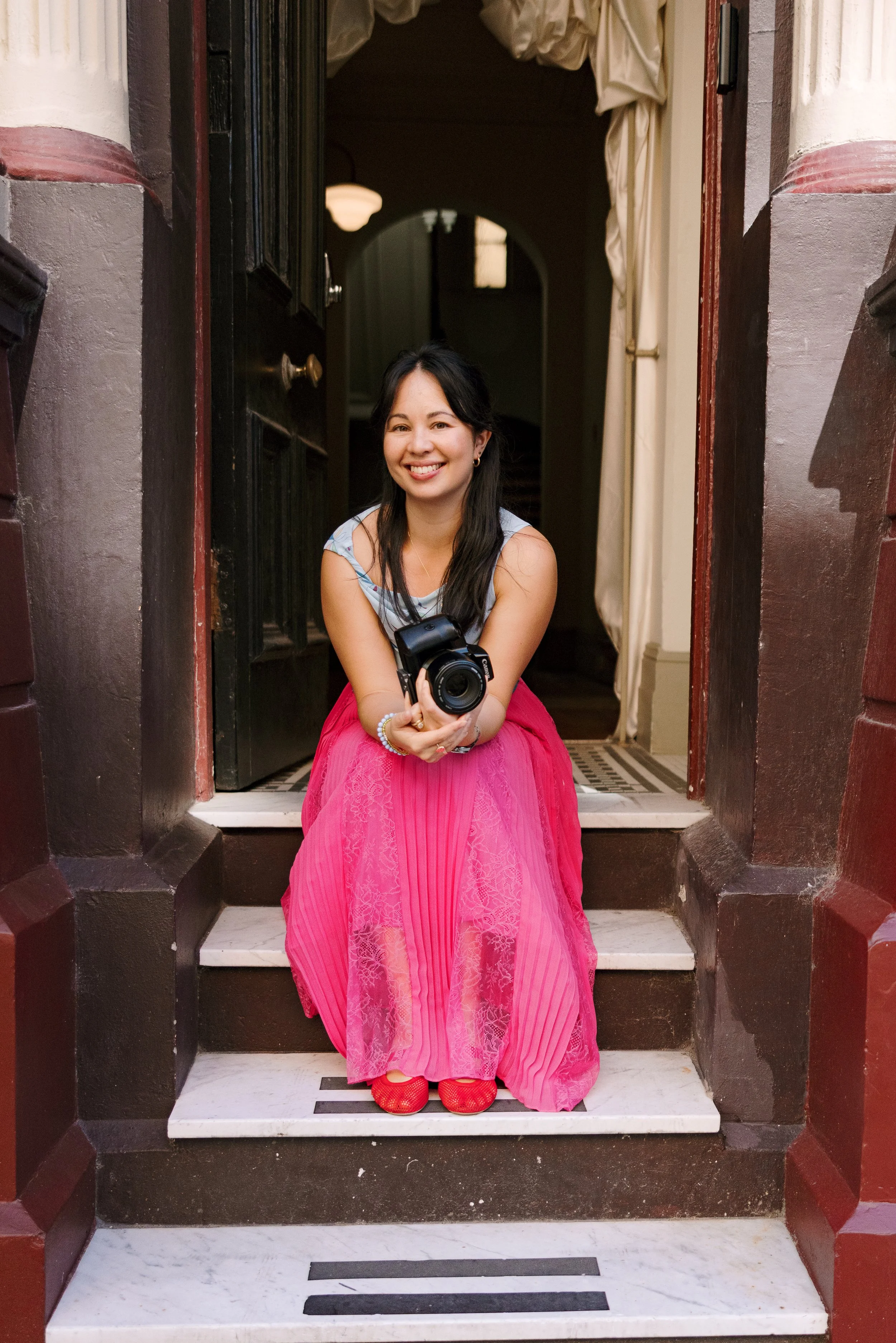 A woman with dark hair, wearing a striped sleeveless top, a long pink skirt, and red shoes, sitting on the stairs at the entrance of a building with a camera in her hands, smiling at the camera.