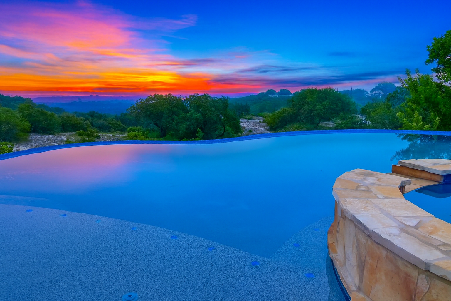Infinity pool with stone edge overlooking a lush green landscape and cloudy sky at sunset.