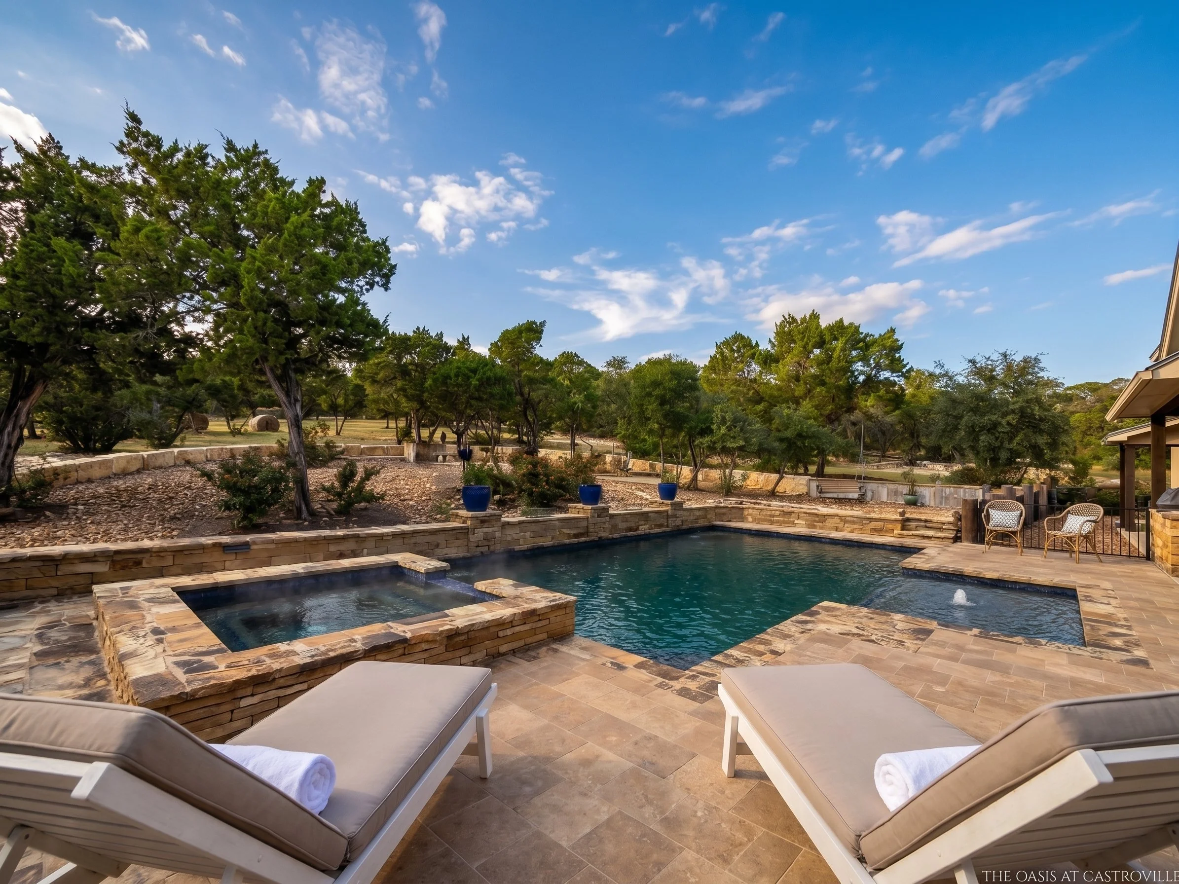 Outdoor swimming pool with a hot tub and waterfall feature surrounded by a stone patio, with trees and blue sky in the background.