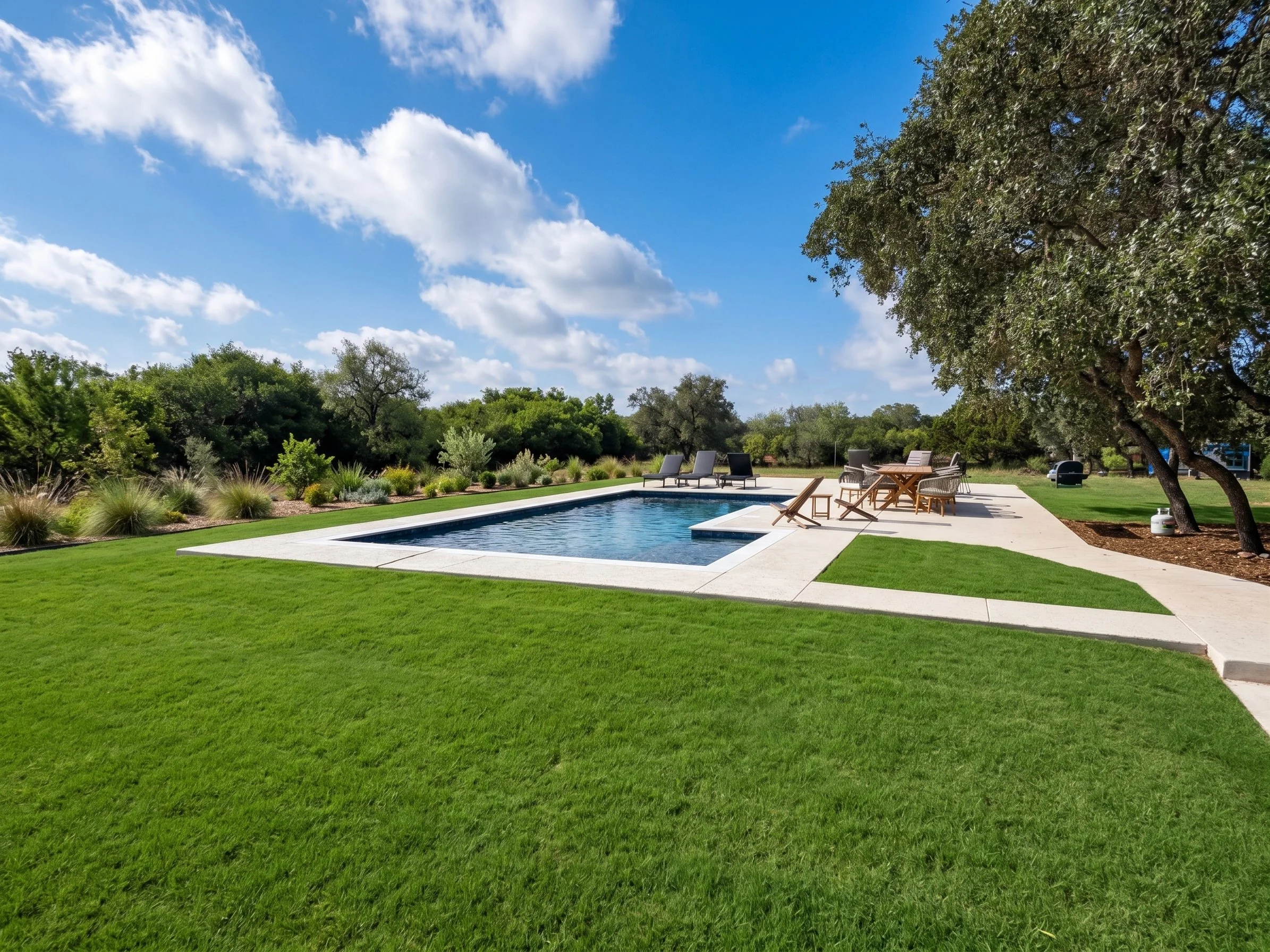 A backyard scene with a swimming pool, outdoor furniture including lounge chairs and a table with chairs, surrounded by lush green grass, trees, and bushes with a partly cloudy blue sky overhead.