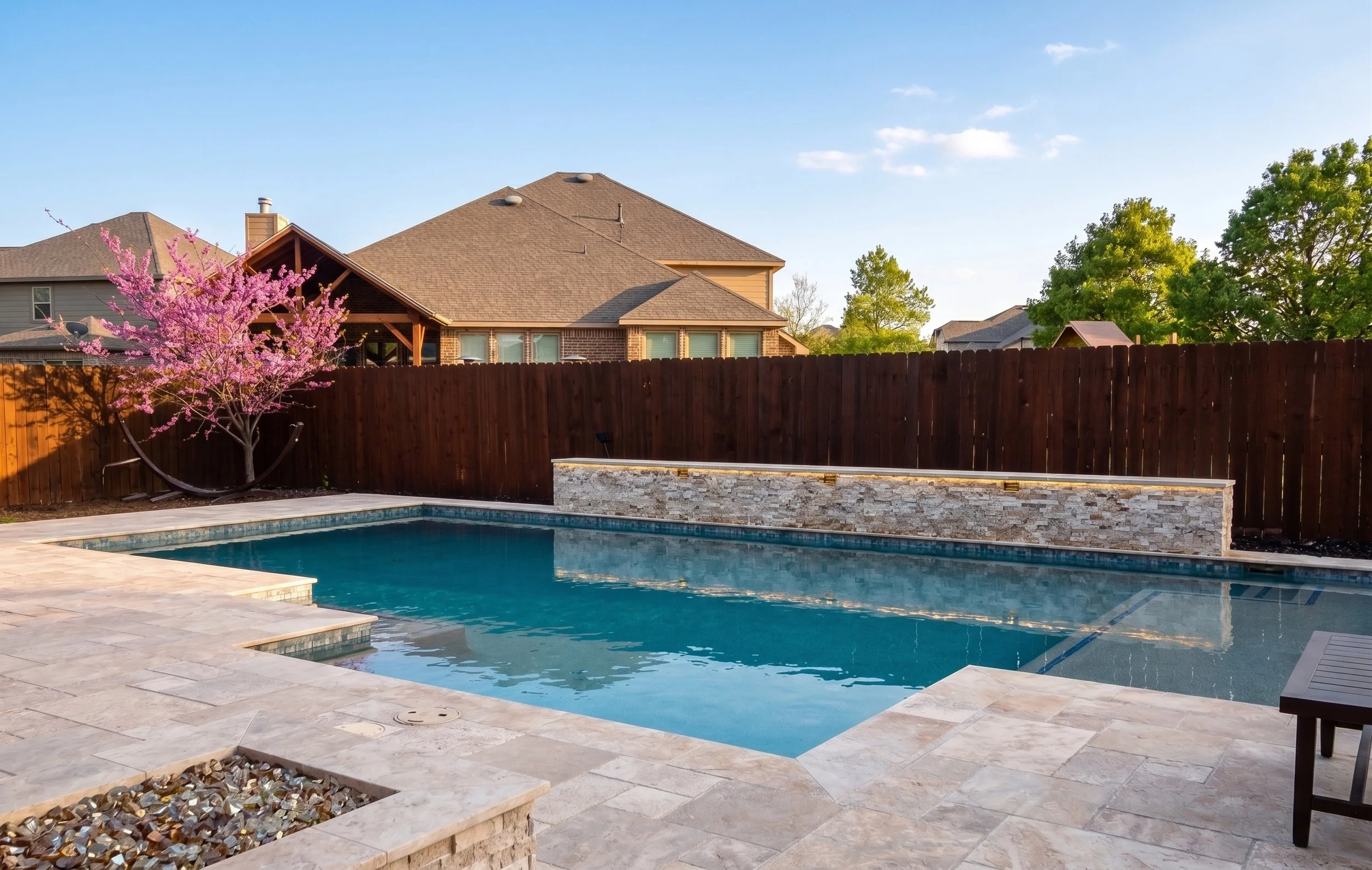A backyard swimming pool with a stone deck, a low stone wall on the far side, a redwood fence, and a tree with pink blossoms on the left side.