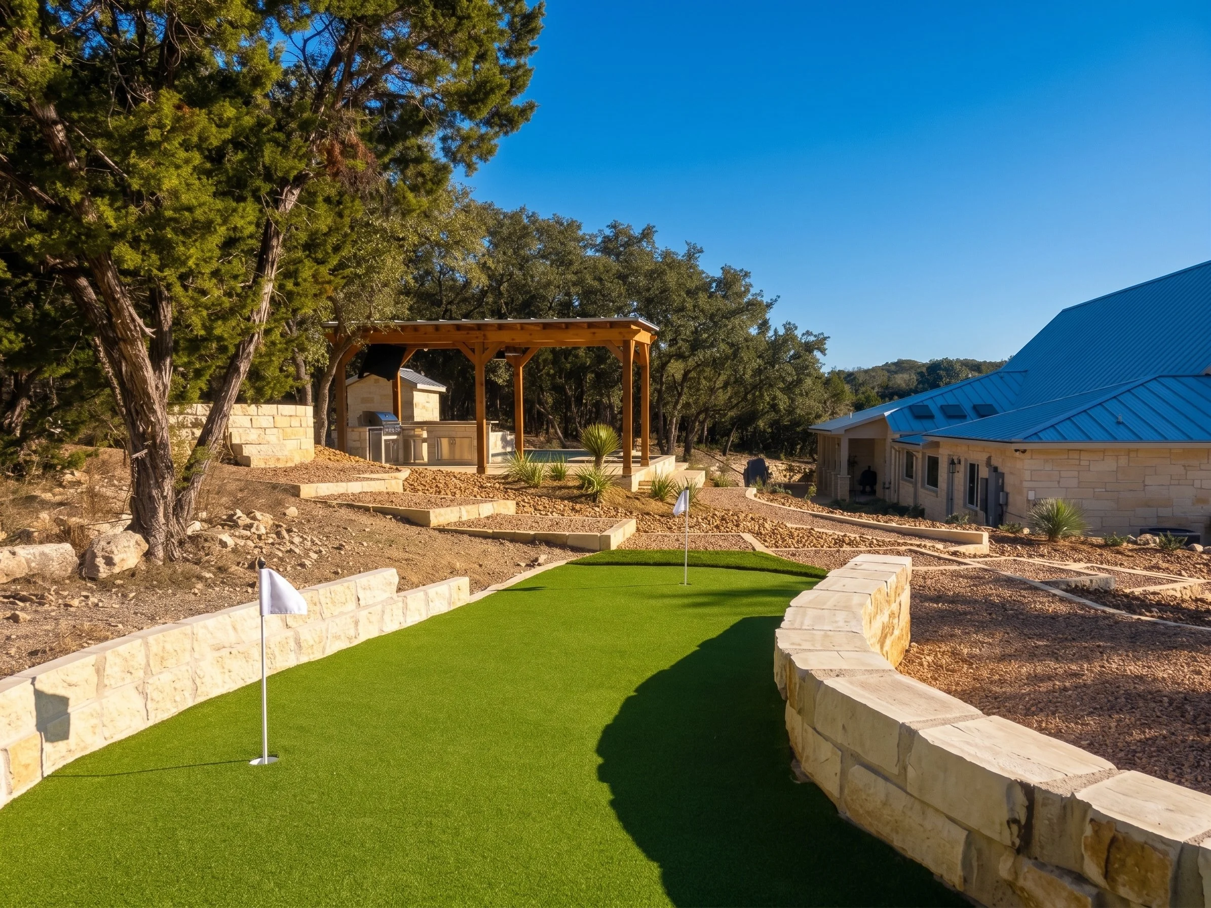 View of a backyard with a putting green, stone pathways, and landscaped plants, with a large house and trees in the background.