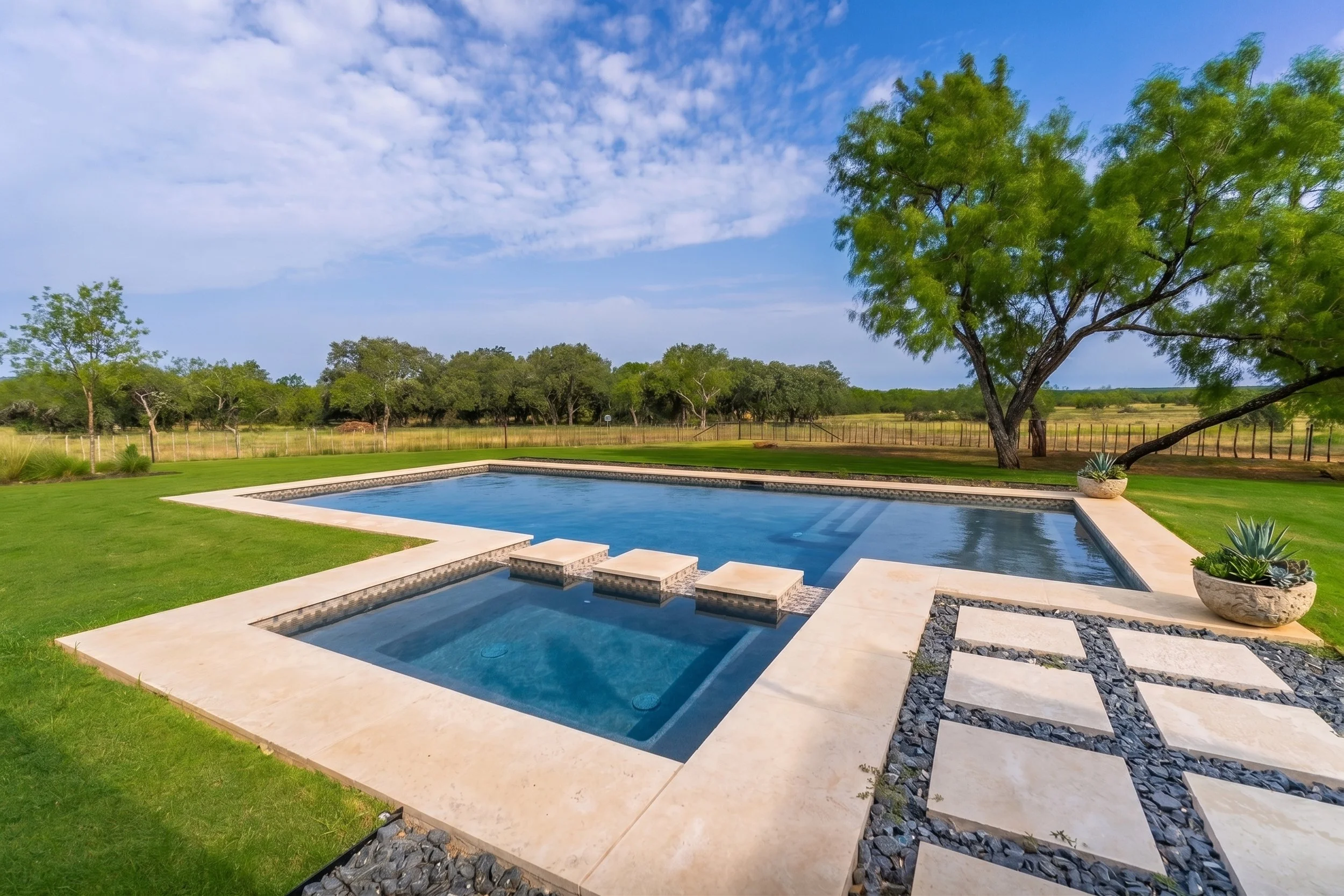 A backyard with a kidney-shaped swimming pool, poolside chairs, a large green tree, and open grassy fields in the distance under a partly cloudy sky.
