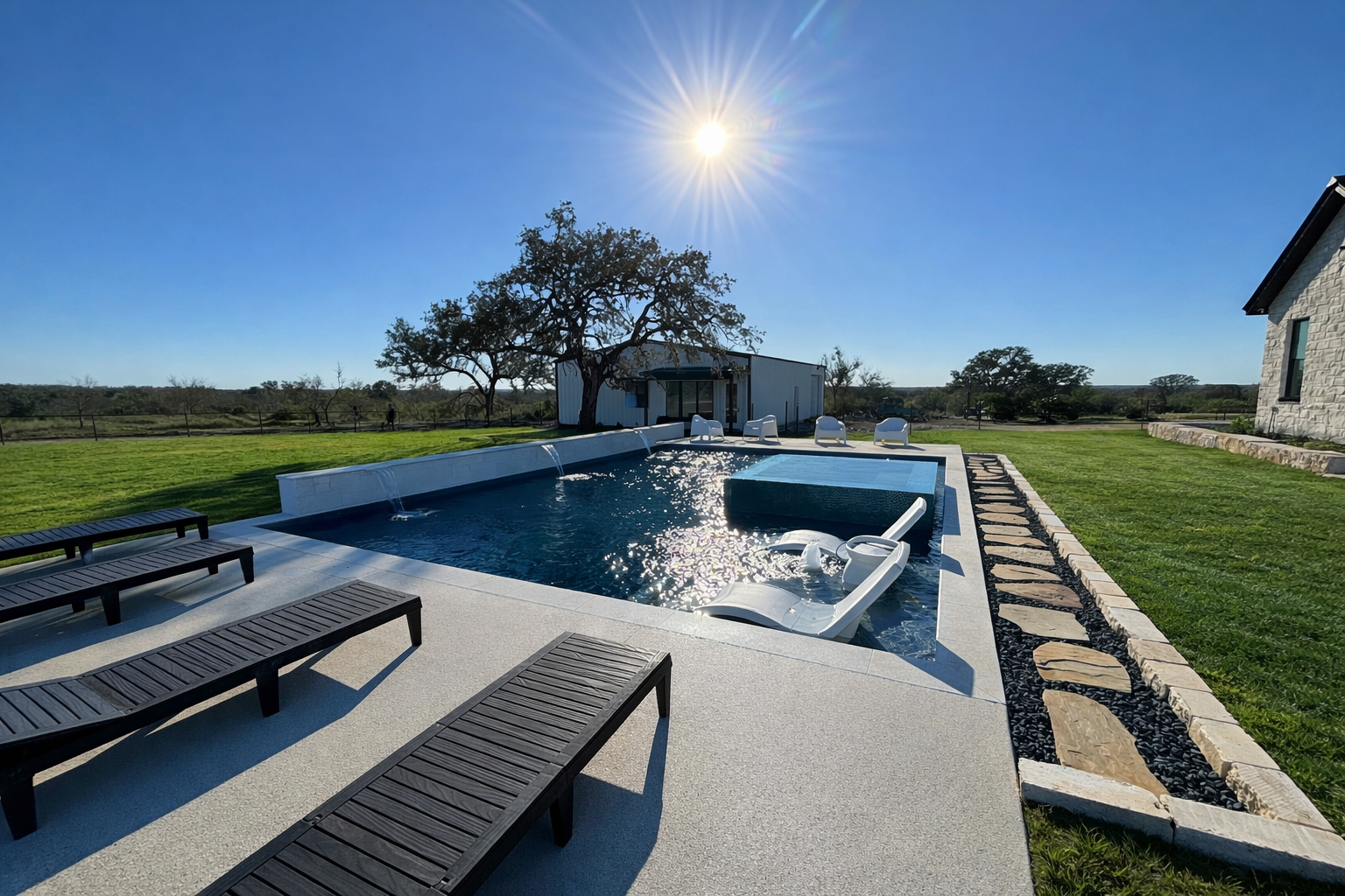 An outdoor swimming pool with lounge chairs, a grassy yard, and a tree with a shed in the background under a bright sun in a rural setting.