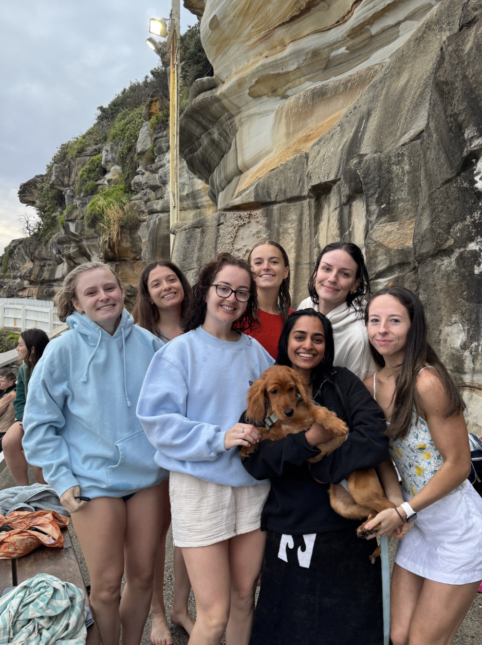 Group of seven young women and a dog standing on a beach with rock formations in the background.