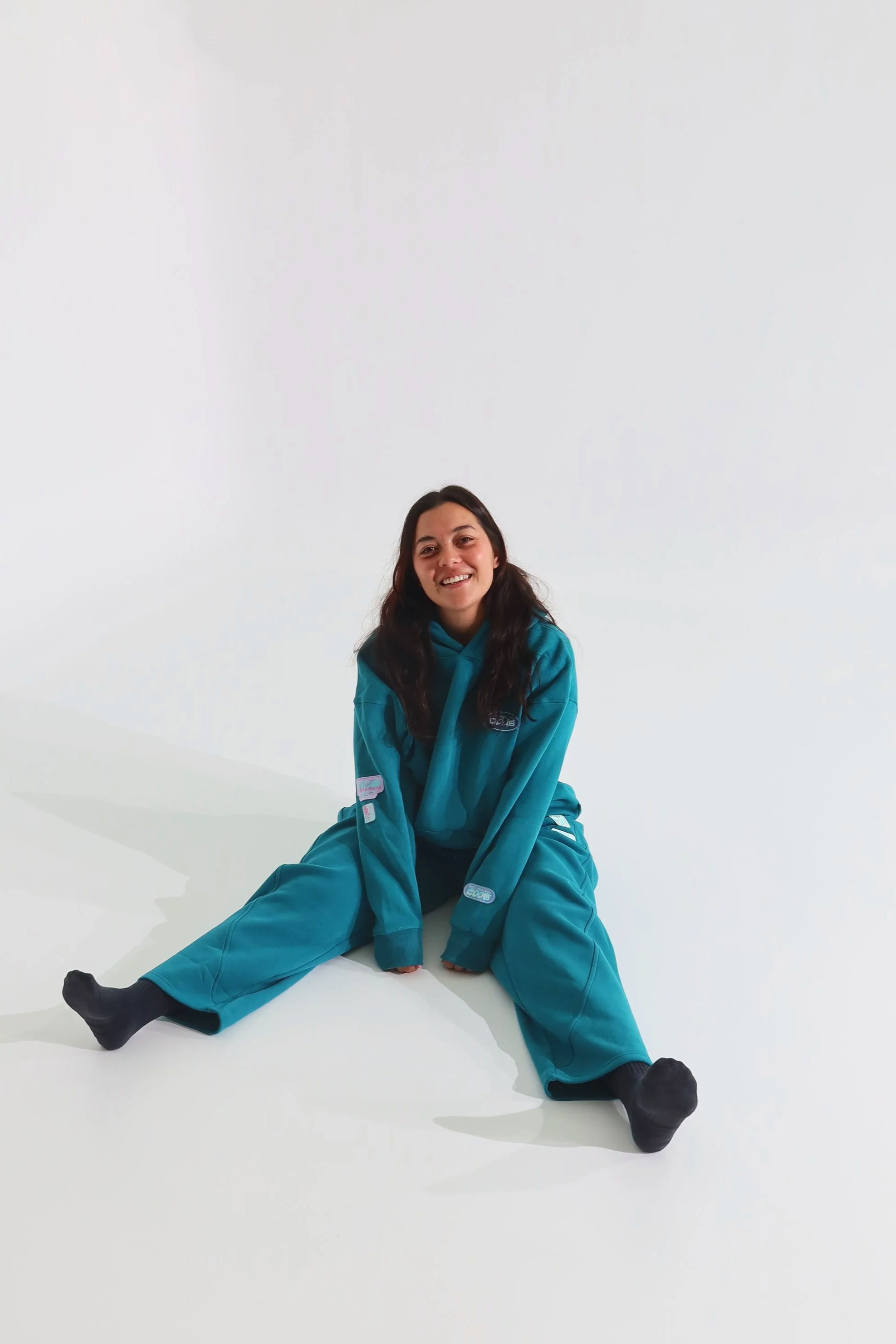A woman sitting on the floor with legs spread apart, smiling, wearing a teal hoodie and matching teal baggy pants, against a plain white background.